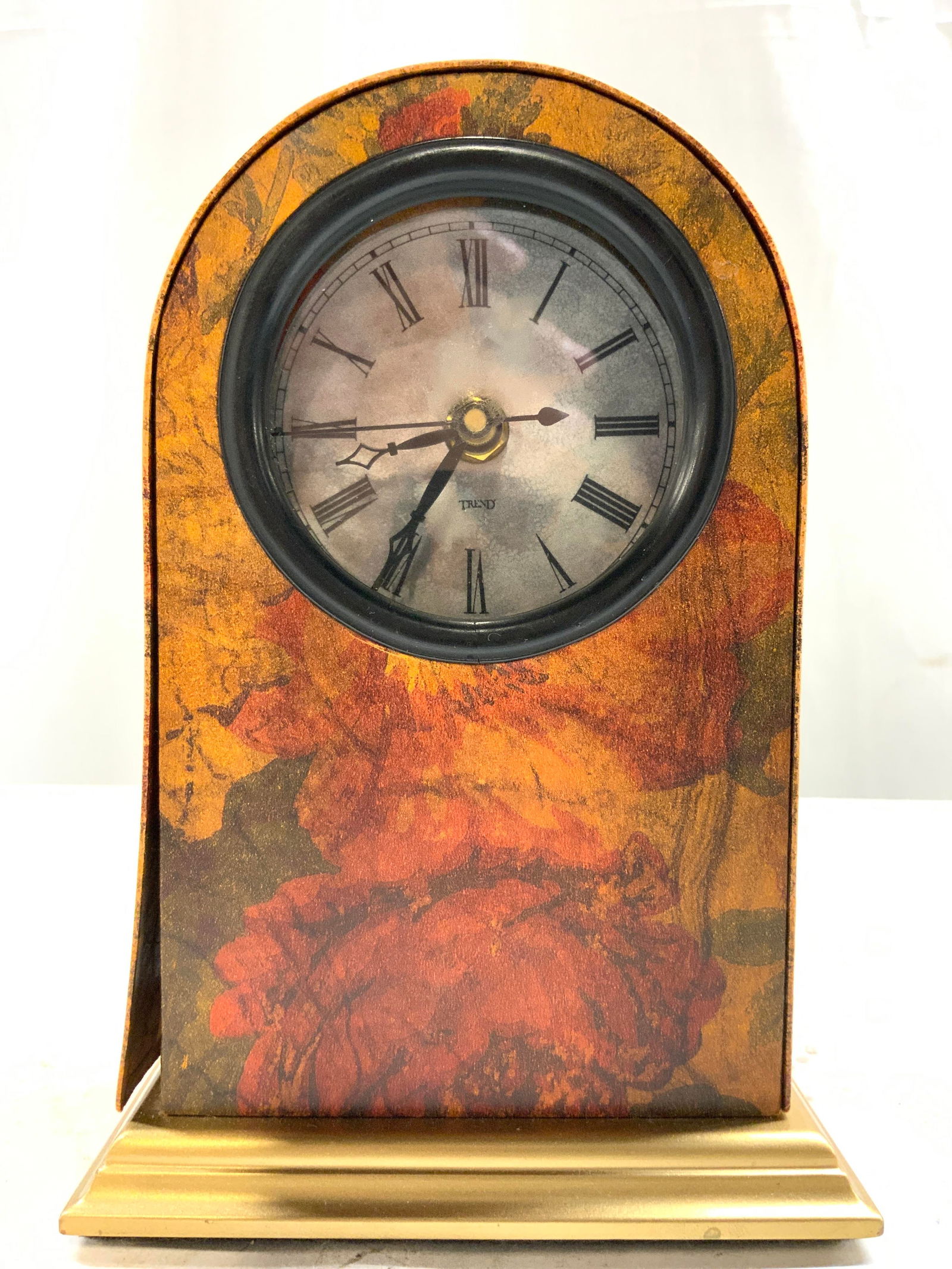 TREND Floral Detailed Arched Mantel Clock: Clock face reads TREND. Clock has multi toned floral detail. Piece has black toned Roman numerals and hands. Clock has gilt metal base. Mantel clock measures approximately 7.25 inches tall 5 inches wi