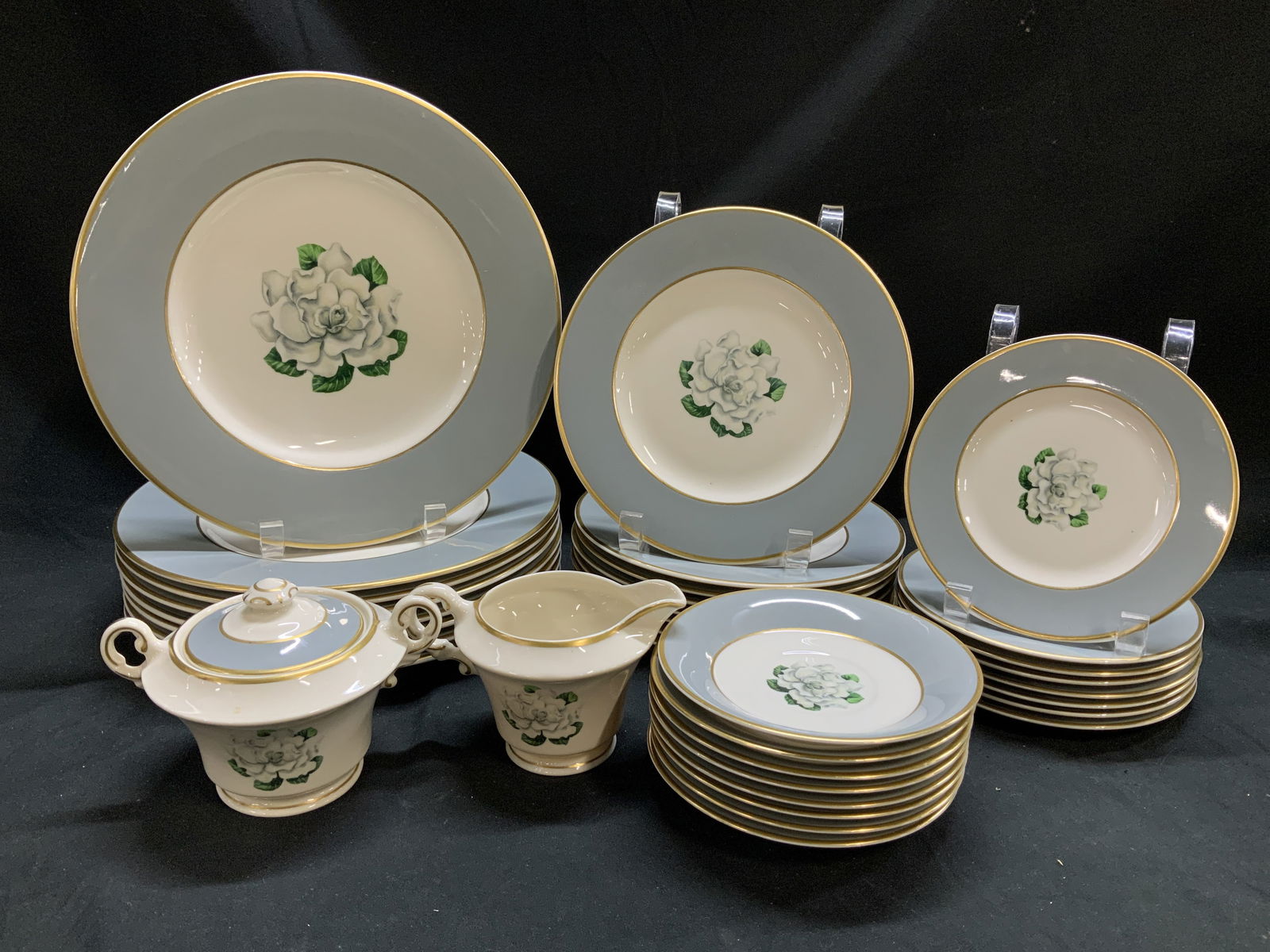 Lot 34 Avalon Syracuse China Floral Tableware: Lot includes, lidded sugar bowl, creamer, 8 saucers, 8 appetizer plates, 8 desert plates & 8 dinner plates. Pieces are porcelain material & feature a white flower w a gray band & gold trim. Surface we