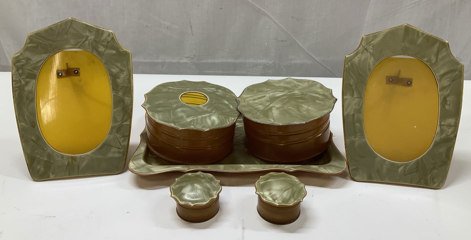 Lot 7 Vntg Green & Amber Celluloid Dresser Set: Possible circa 1930s. Dresser set includes a powder box, hair receiver, tray, 2 picture frames & 2 round trinket boxes. Pieces are celluloid material & feature amber brown bases with a pearlescent lig