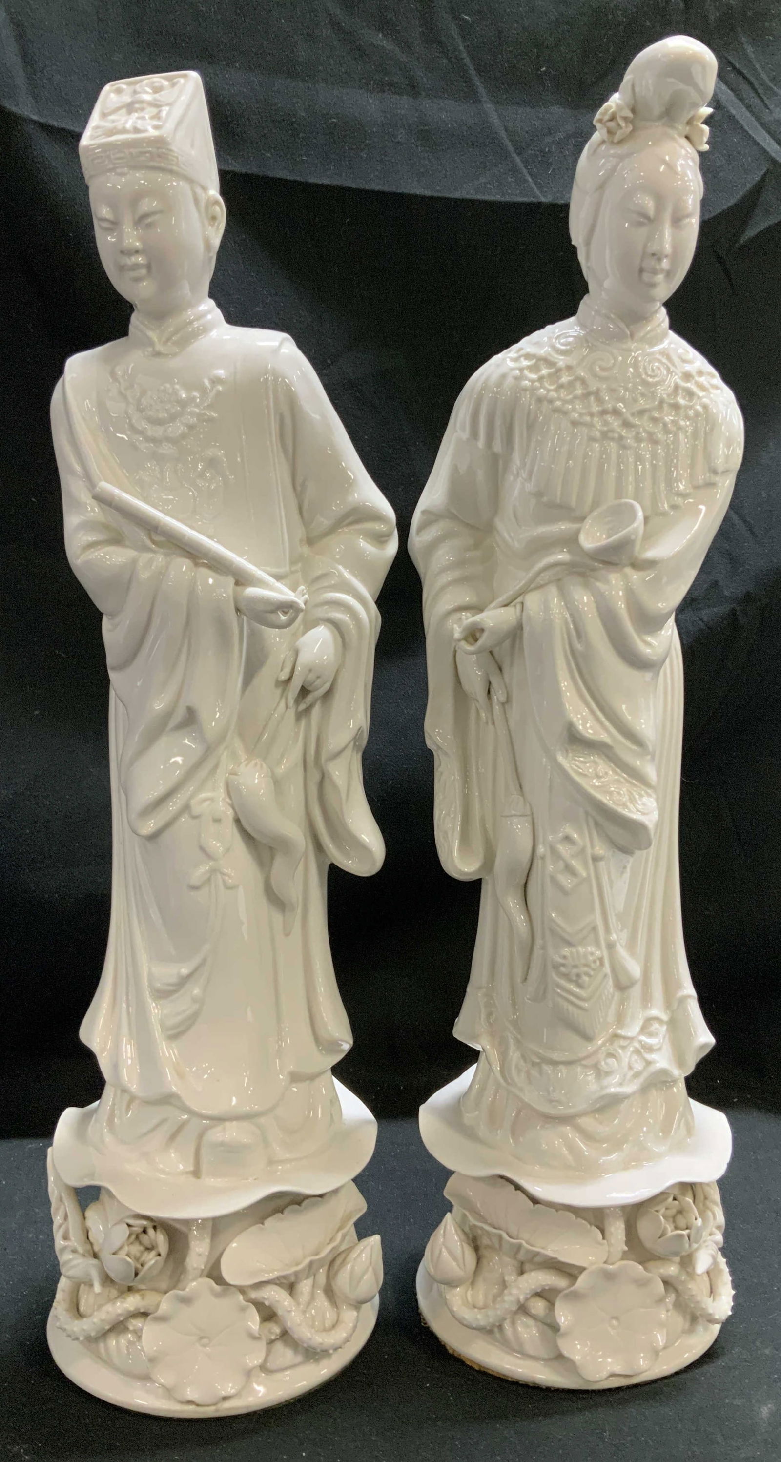 Lot 2 Blanc De Chine Asian Man & Women Figurines: Male figurine is wearing a traditional scholars robe & a square hat. Female is dressed in a robe w intricate details. Both figures are standing on pedestal bases adorned with lotus motifs. Blance de c