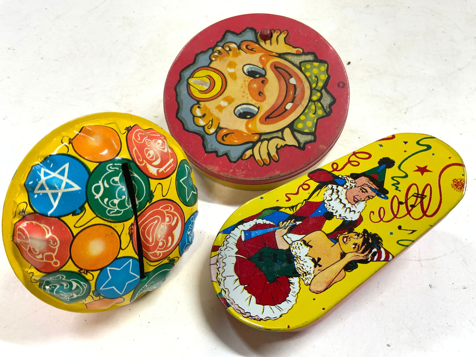 Lot 3 Vntg Tin Lithographed Noisemakers: Lot includes 3 Noisemakers. One circular form with image of clown T COHN, 1 with image of balloons & 1 with a male and female figure dressed in holiday attire US METAL TOY CORP. Each noisemaker is vin