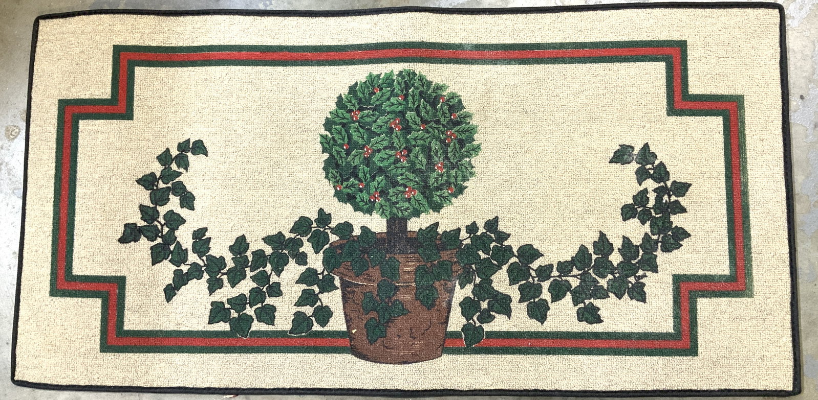 Potted Ivy & Topiary Accent Doormat 39.6in: Doormat features a design of a potted plant with ivy & a holly bush. Features a red & green border & black trim edge. Appears to be synthetic material. No visible signature or markings. Surface wear c