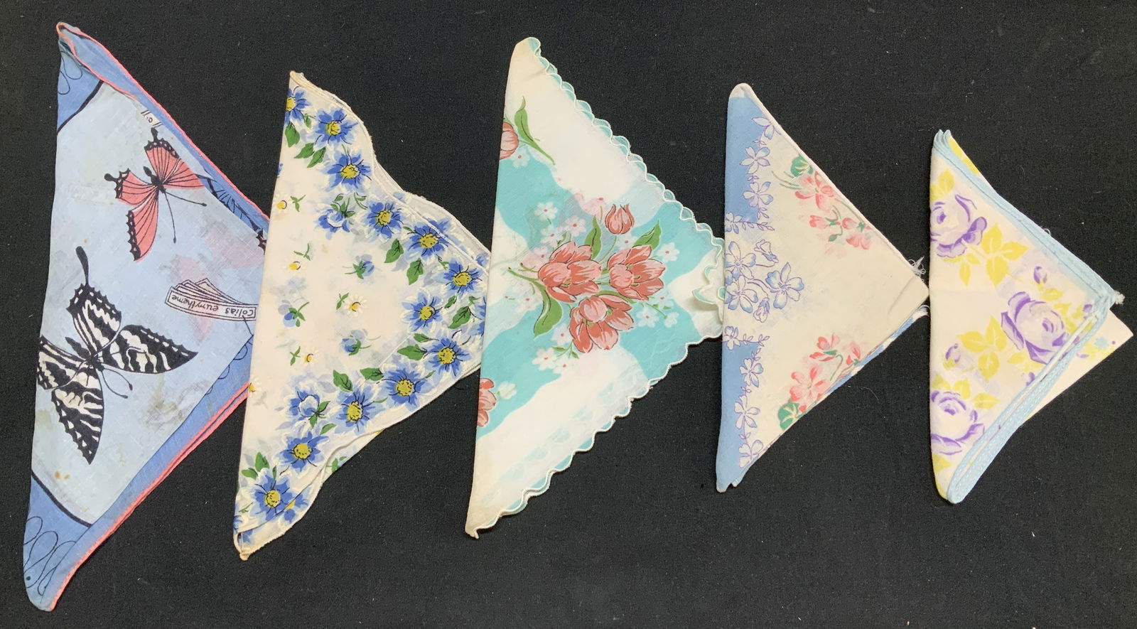 Lot 5 Assorted Floral & Butterfly Handkerchiefs: Handkerchiefs appear to be cotton material. They feature floral pattern in shades of pink, green, blue, yellow & purple. 1 handkerchief features a scalloped edge. 1 handkerchief features a Butterfly p