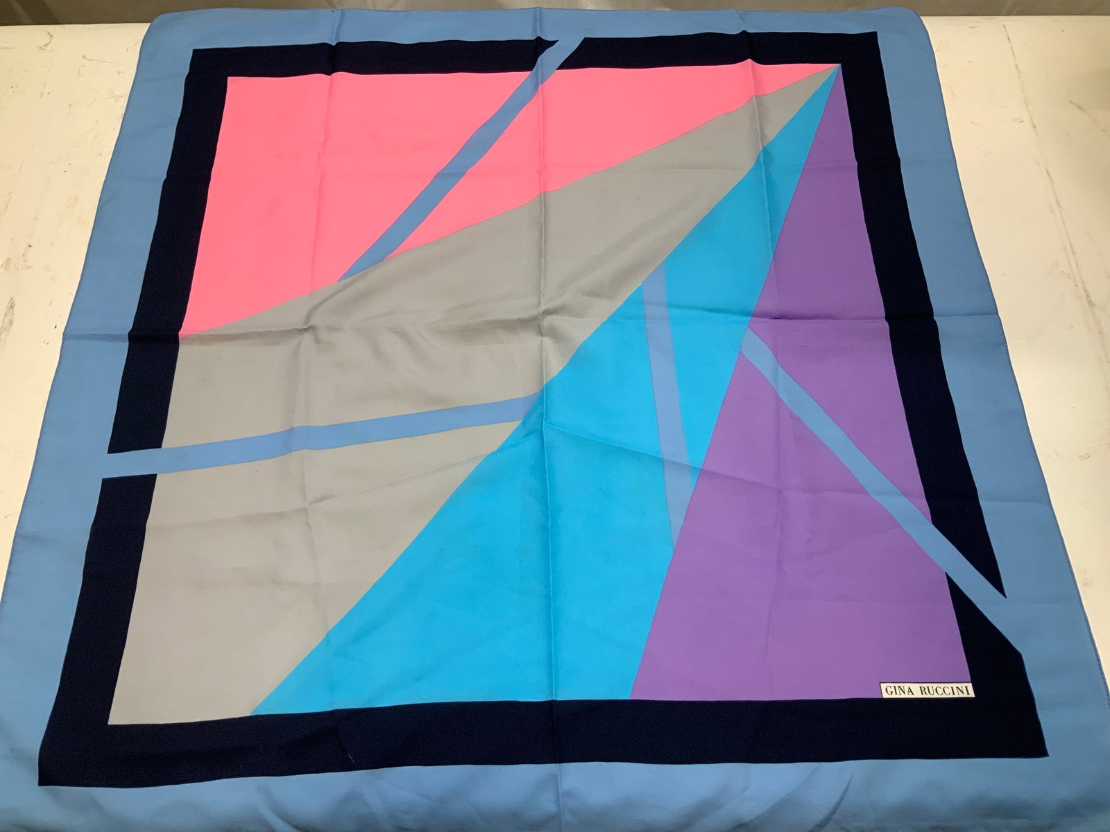 Vntg Gina Ruccini Geometric Print Scarf: Scarf features a geometric design with shades of pink, blue, purple & grey. Scarf appears to be possible polyester material. Bottom right reads, GINA RUCCINI. Surface wear consistent to age, use & exp