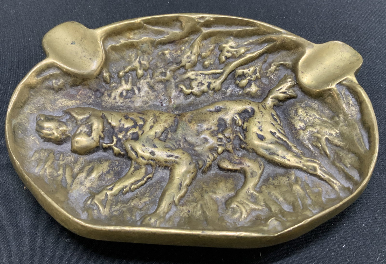 Vintage Hunting Dog Brass Ashtray: Ashtray features a bas-relief of a hunting dog in a field scene. Ashtray is brass material. Textured details & 2 indentations. Surface wear consistent to age, use & exposure. Measurements: length 5.5i