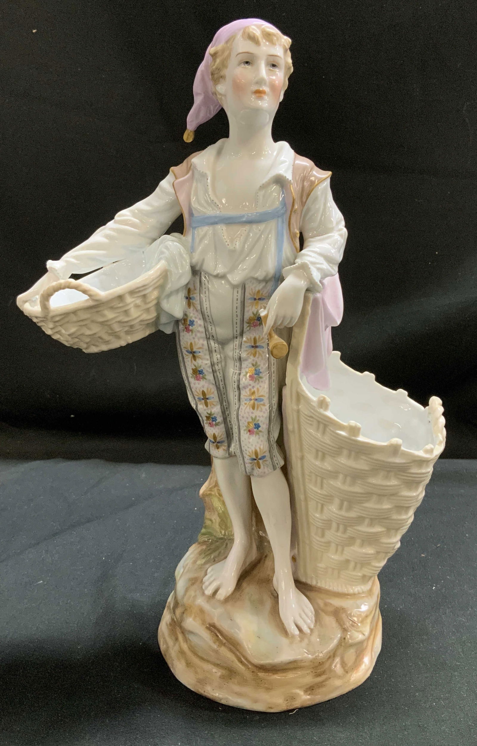 Signed Vntg Barefoot Boy W Baskets Porcelain Fig.: Porcelain figurine depicts a barefoot boy wearing a white shirt, and striped pants with floral embroidery. He is standing on a rocky base. 2 open baskets. Surface wear consistent to age, use & exposur