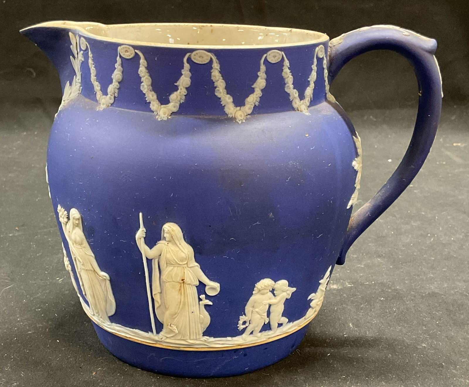 Vtg Wedgwood Blue Jasperware Pitcher: Vintage Wedgwood cobalt blue Jasperware pitcher. White bas relief with classical figured and garland border. Signed. Made in England. Measures approx 7 x 5 x 5.5 inches. Some wear including chip under