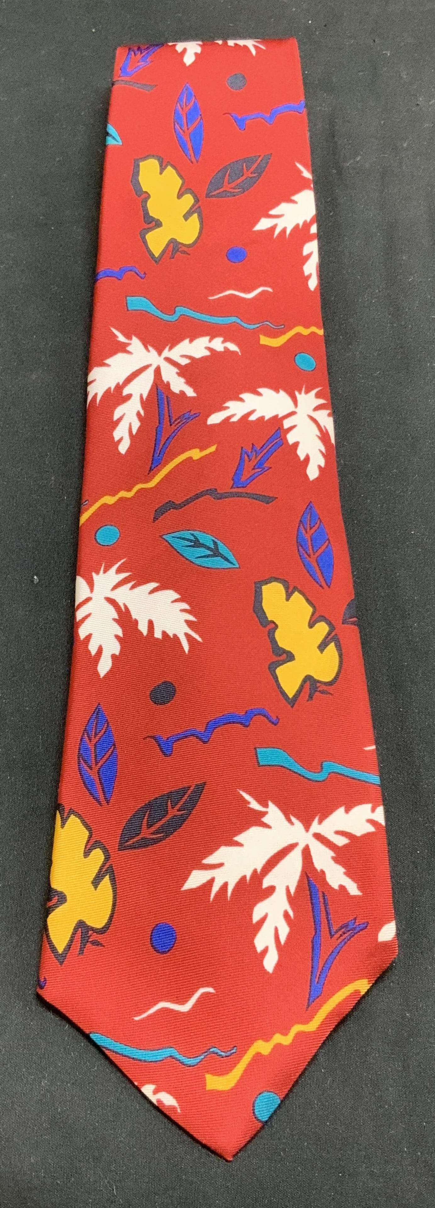 Cacharel Hand Printed Tropical Silk Necktie ITALY: Red necktie with a tropical pattern featuring white, yellow, blue & black tones. Hand printed. Signed & marked. Reads, CACHAREL. PARIS. HAND PRINTED IN ITALY. ALL SILK. Surface wear consistent to age,