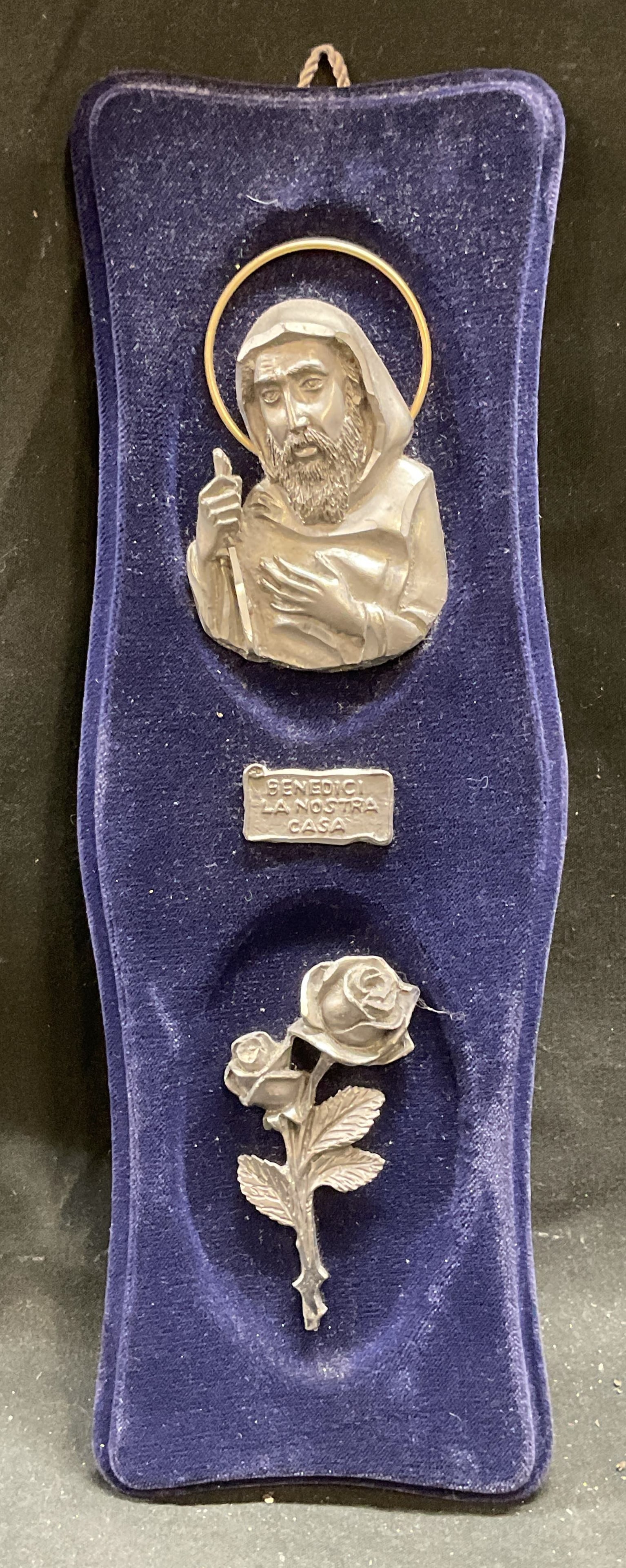 Bless Our Home Pewter Religious Plaque: Pewter relief plaque mounted in blue velvet. Benedici La Nostra Casa. Bless our home. Image of a saint and roses. Measures approx 10.5 x 3.5 inches. Some wear consistent with exposure. Pewter plaque,