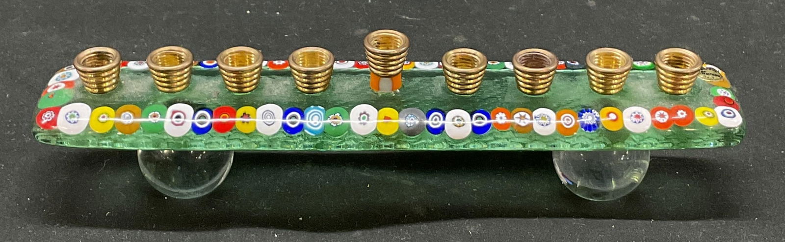 Judaica Glass Patterned Menorah: Judaica glass patterned Menorah. Made of translucent glass fused base. Round multi colored pebble pattern embedded along edges. Brass toned metal candle holders on top surface of glass base. Clear sph