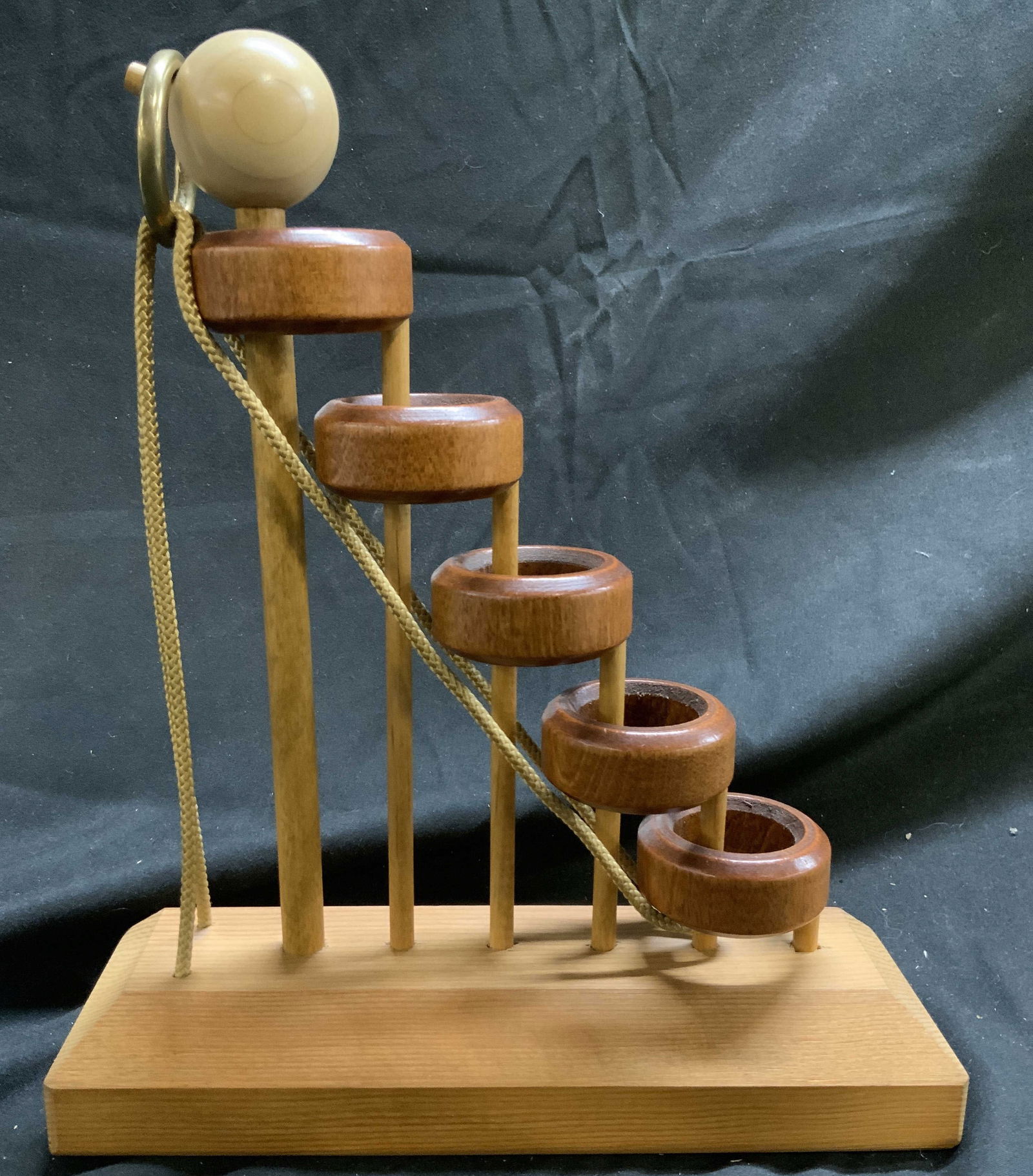 LUMBERJACK TOYS Type M Wood Stump Puzzle 1979: Underside is marked TYM M Lumberjack Toys 1979 San Francisco 94110. Puzzle is wooden. Piece has 5 ring structures 1 string and 1 spherical form structure. Base in tan toned and wooden. Puzzle toy Brai