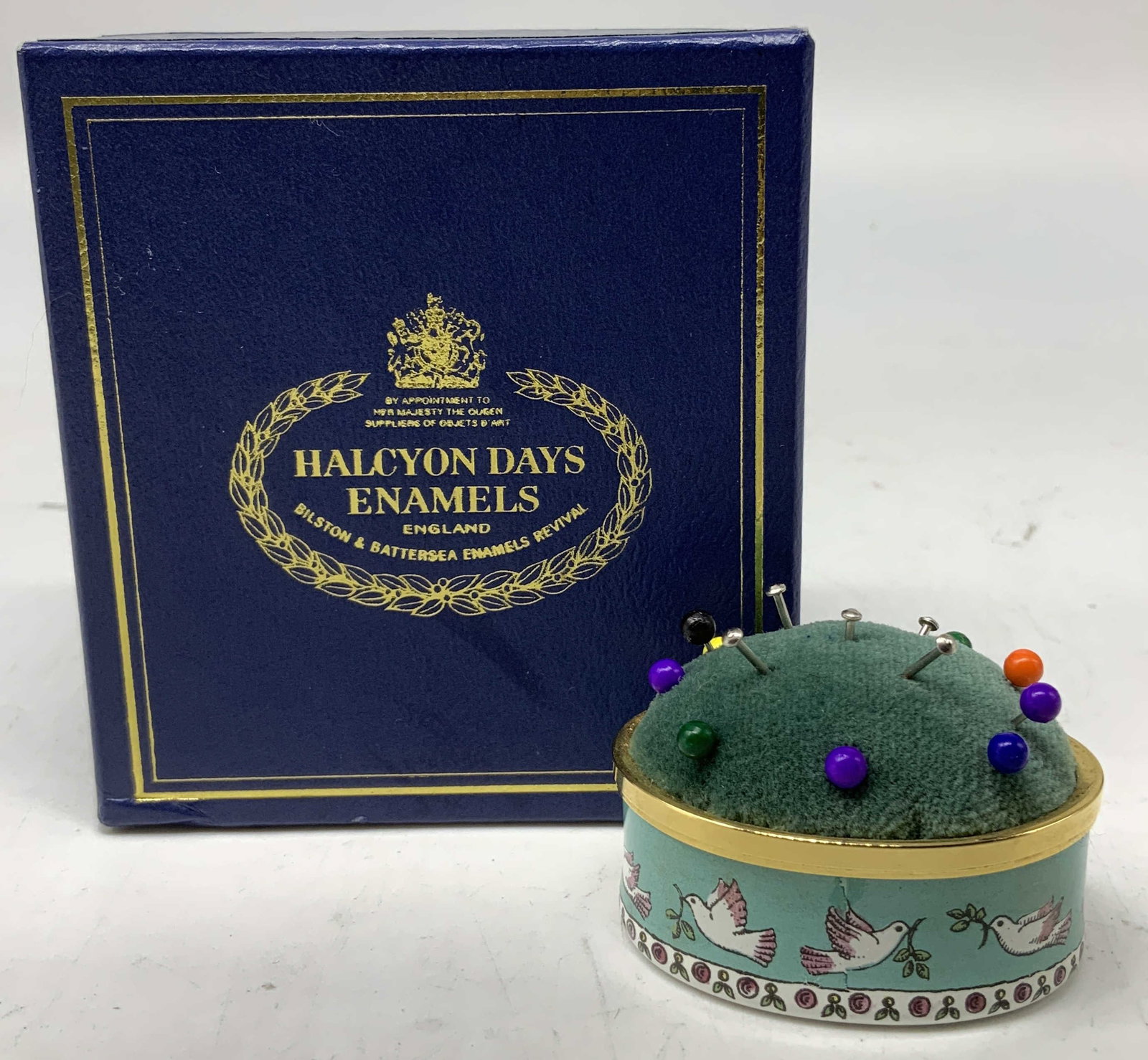 Halcyon Days Enamel Doves Pin Cushion W Box: Halcyon Days enamel pin cushion. Base features an illustration of doves carrying branches. Green pin cushion on top. Comes with box & story of Halcyon Days Enamels. Surface wear consistent to age, use