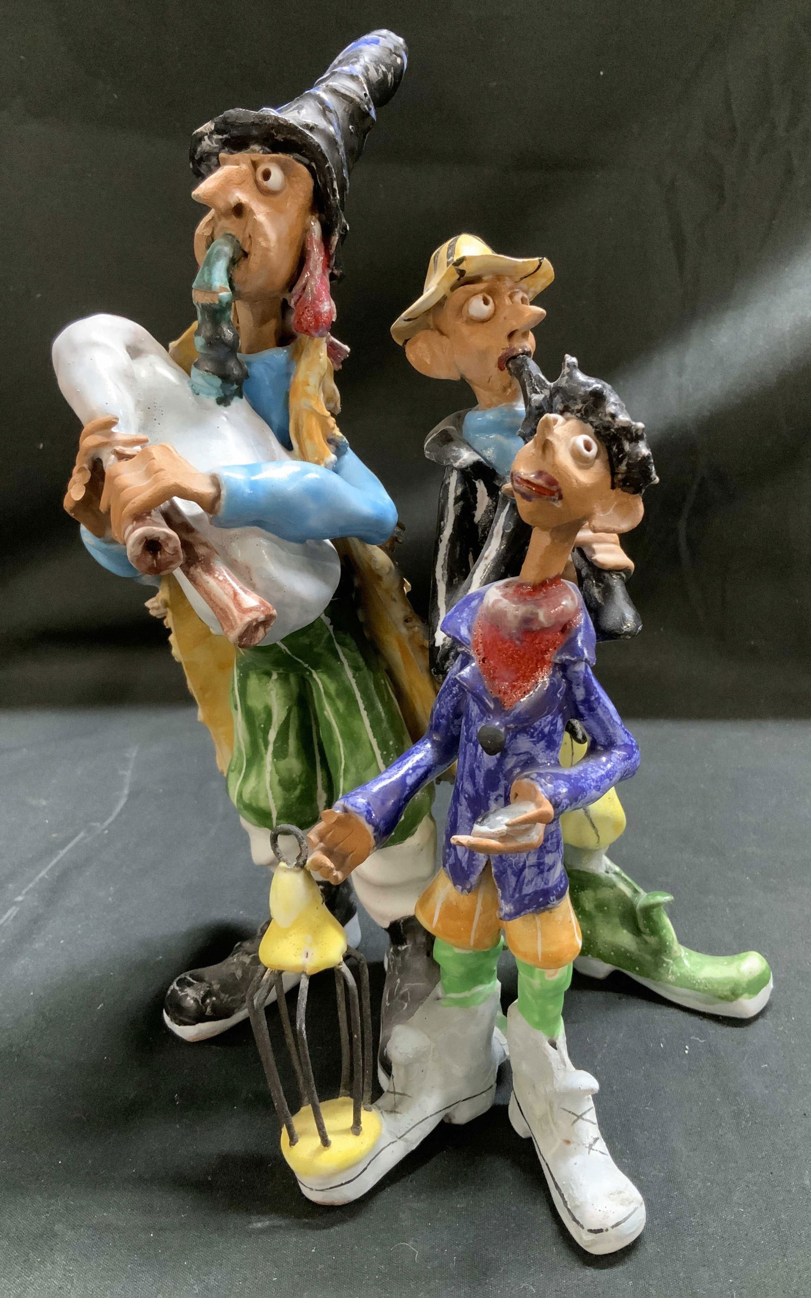 Whimsical Trio Street Musicians Ceramic Figurines: Hand-painted figurines depicting 3 street musicians performing together. Each figurine features elongated limbs & are wearing quirky clothing. Tallest fig is playing a bagpipe like instrument. Surface