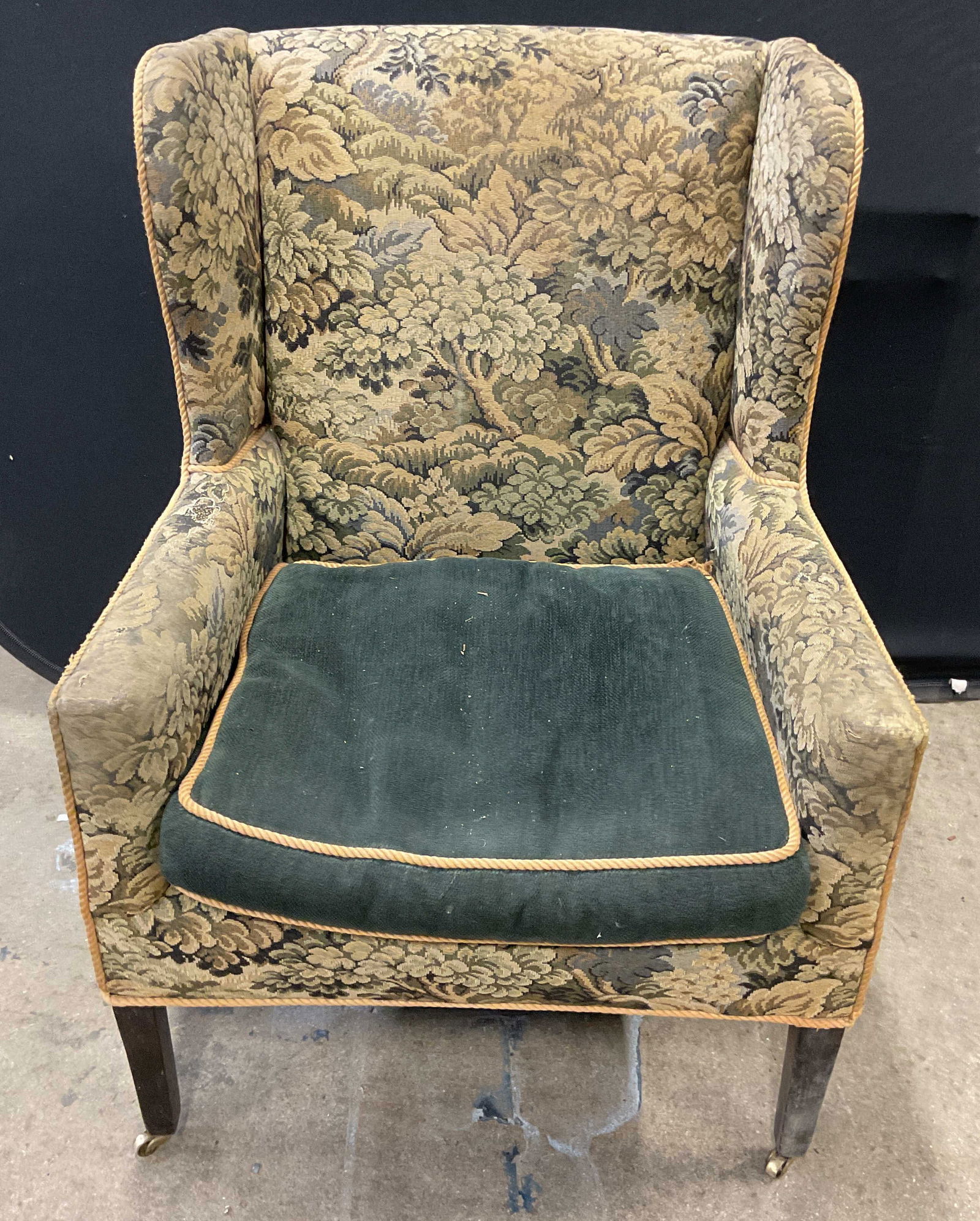 Vintage Green Upholstered Wingback Armchair: Vintage wingback green botanical upholstered armchair with solid green fabric cushion. Wheeled legs. Measure approx 30 x 28 x 41.5 inches Wear including discoloration and tears. Wingback chair, uphols