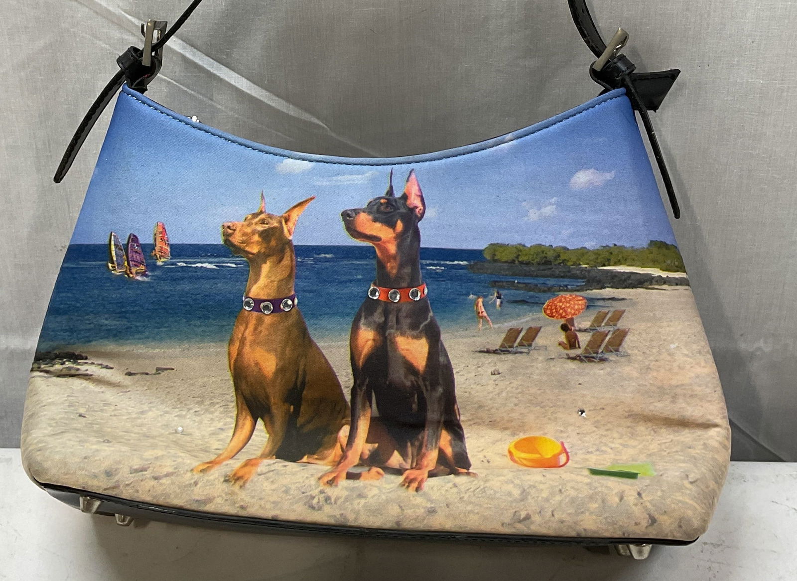 Vintage Doberman Beach Shoulder Bag: Vintage Doberman beach shoulder bag. Feels like cloth. Curved top edge with metallic feet on underside. Depicts beach scene with two Doberman dogs. Studded accents. Faux leather buckle shoulder strap
