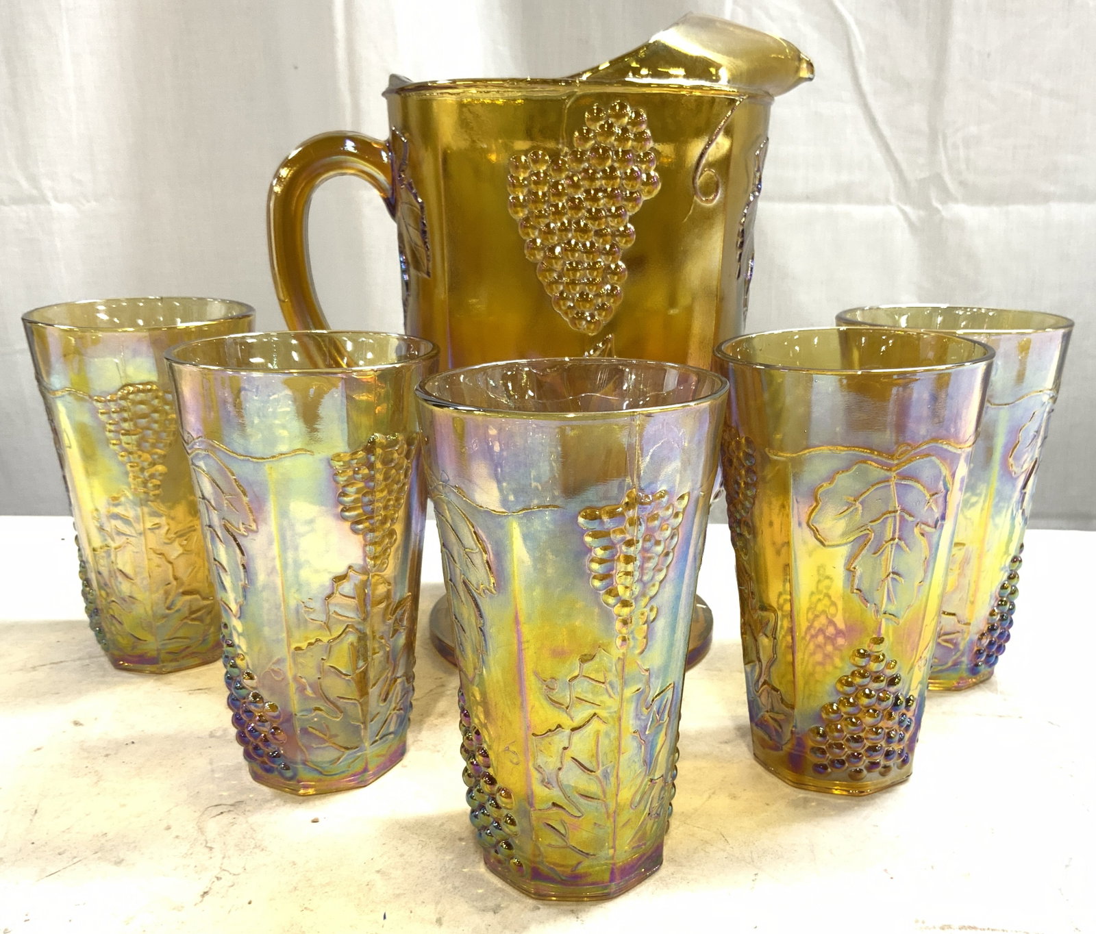 Set 6 Vtg Iridescent Carnival Glass Pitcher & Cups: Lot includes 1 pitcher & 5 cups. Each piece is gold toned iridescent carnival glass. Each vessel has beveled grape and leaf detailing. Pitcher is footed. Set appears to be vintage. Pitcher measures ap