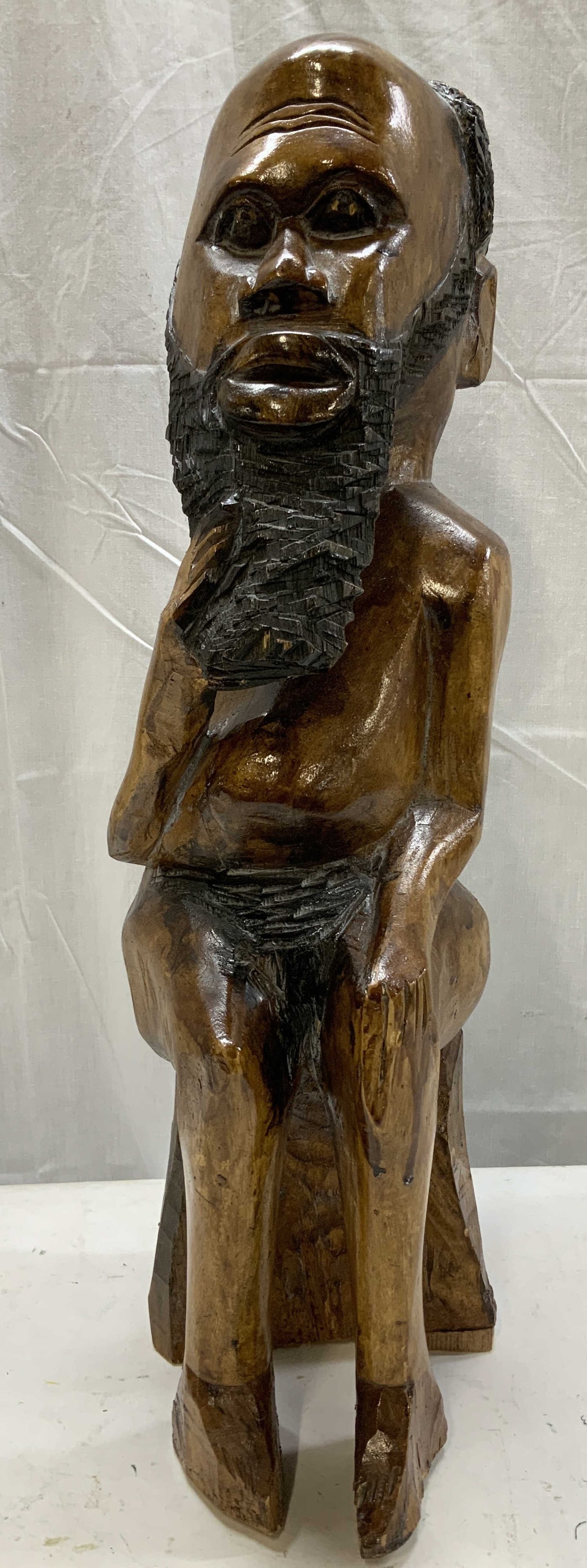 Hand Carved Jamaican Folk Art Style Sculpture: Hand-carved wooden sculpture depicting a seated elder with an elongated head, carved textured beard & facial features. Figure sits on a block-style stool, posed with one hand resting on the knee. Warm