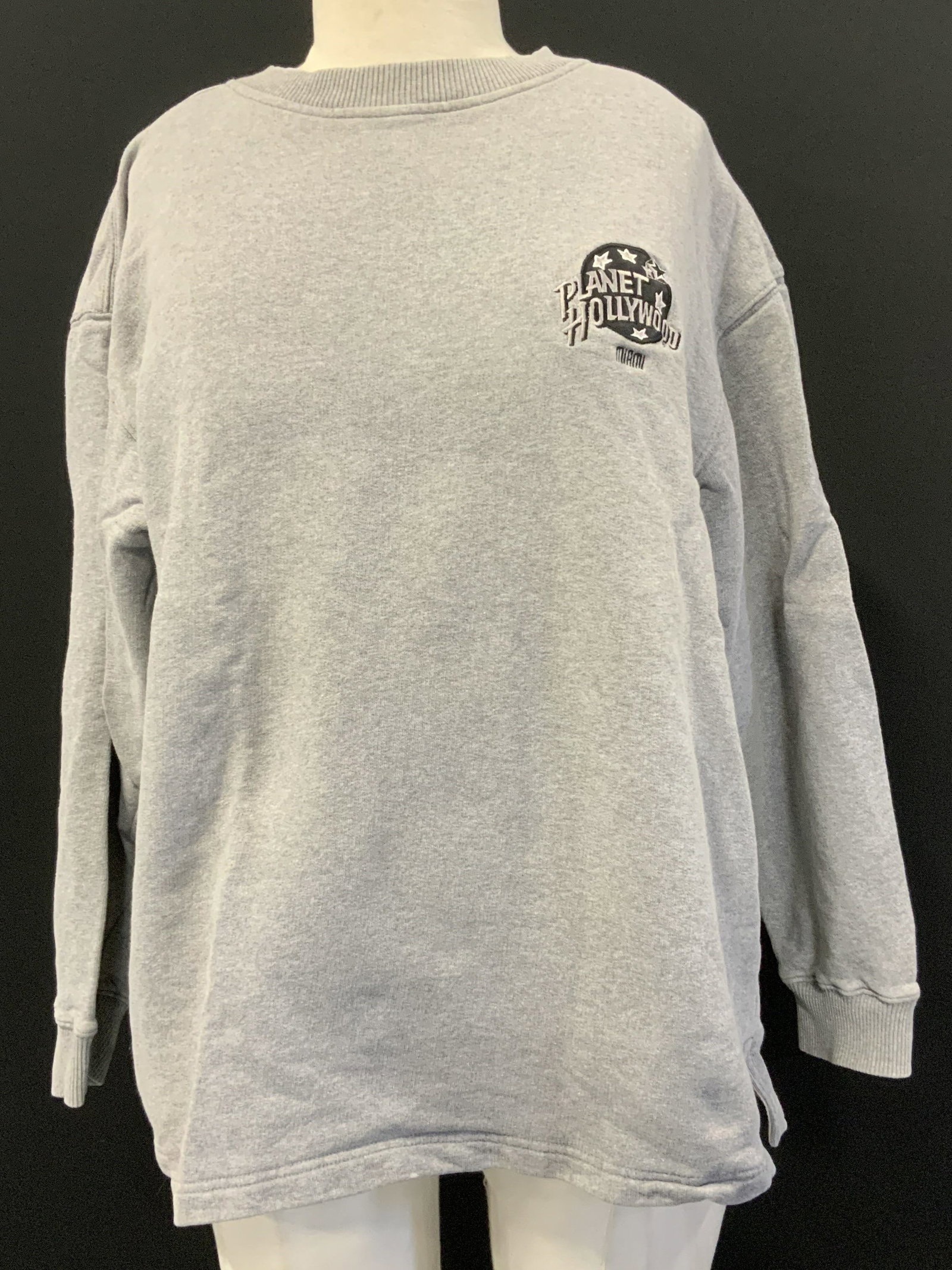 1991 Planet Hollywood Grey Embroidered Crewneck: Crewneck is grey toned & features an embroidered logo. Reads, Planet Hollywood Miami. Logo has white stars. Bottom of crewneck has side slits. Neckline has a ribbed style. Signed & marked. Reads, 1991