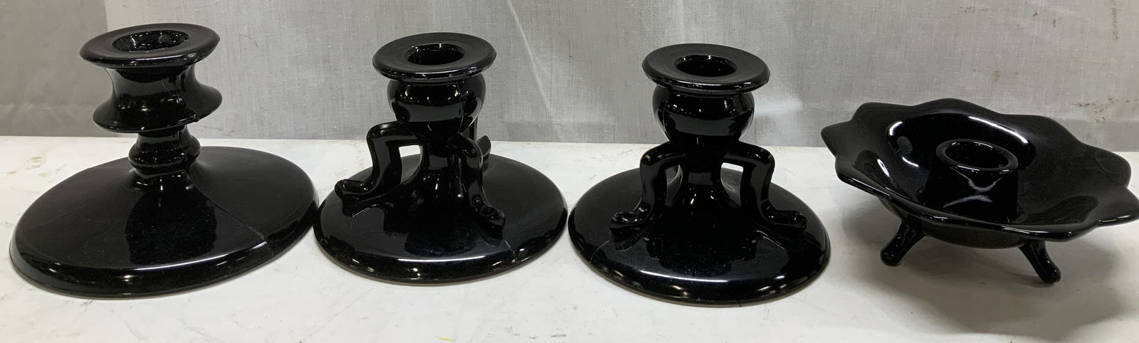 Lot 4 Vntg Assorted Black Glass Candlesticks: Lot includes, 4 assorted candlesticks. Candlesticks are black glass w a glossy finish. 2 candlesticks feature a circular base w 3 feet, 1 candlestick w a spool shaped stem & 1 petal style candlestick.