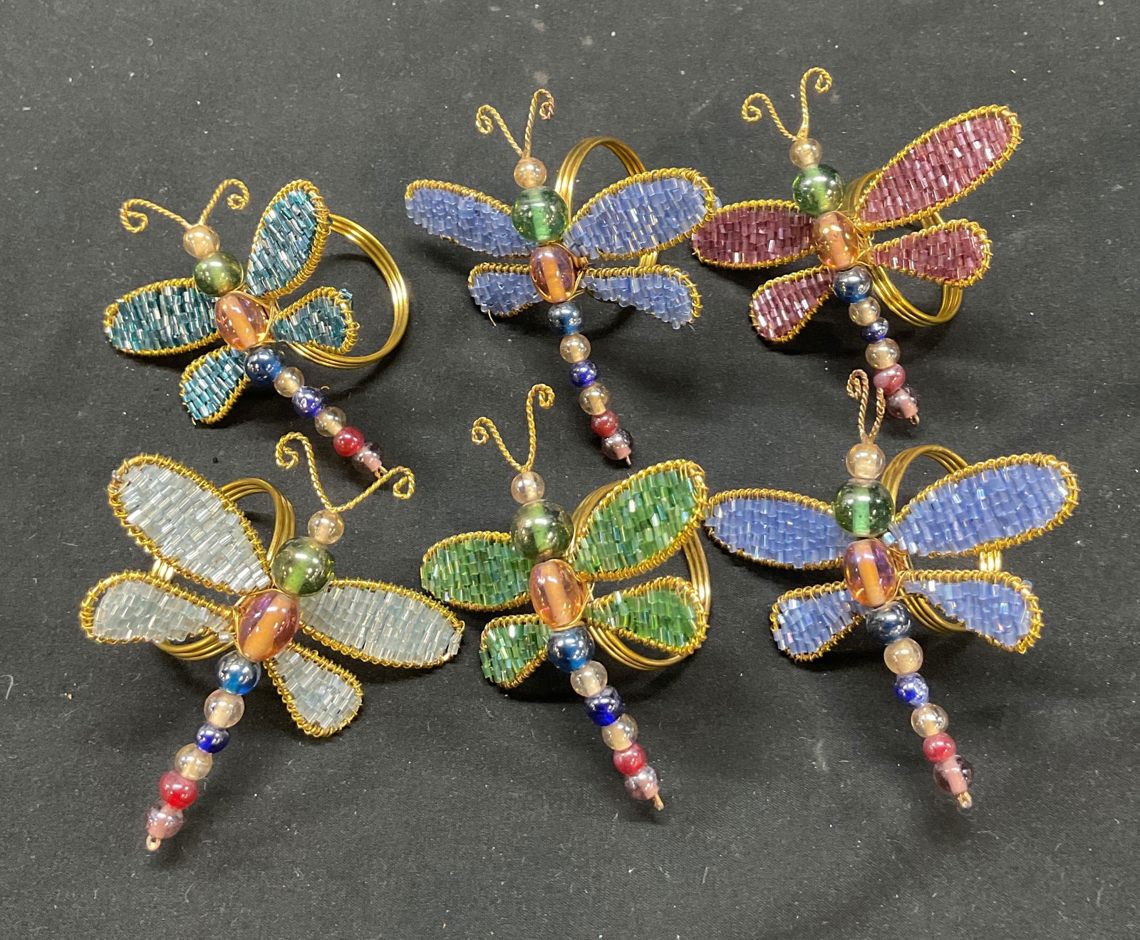 Set 6 Beaded Dragonfly Napkin Rings: Set of 6 beaded dragonfly napkin rings. Feels like metal and glass. Gold toned rings on underside. Dragonfly silhouette with colorful beads. Colors include blue, red, purple, green, and more. Wire gol