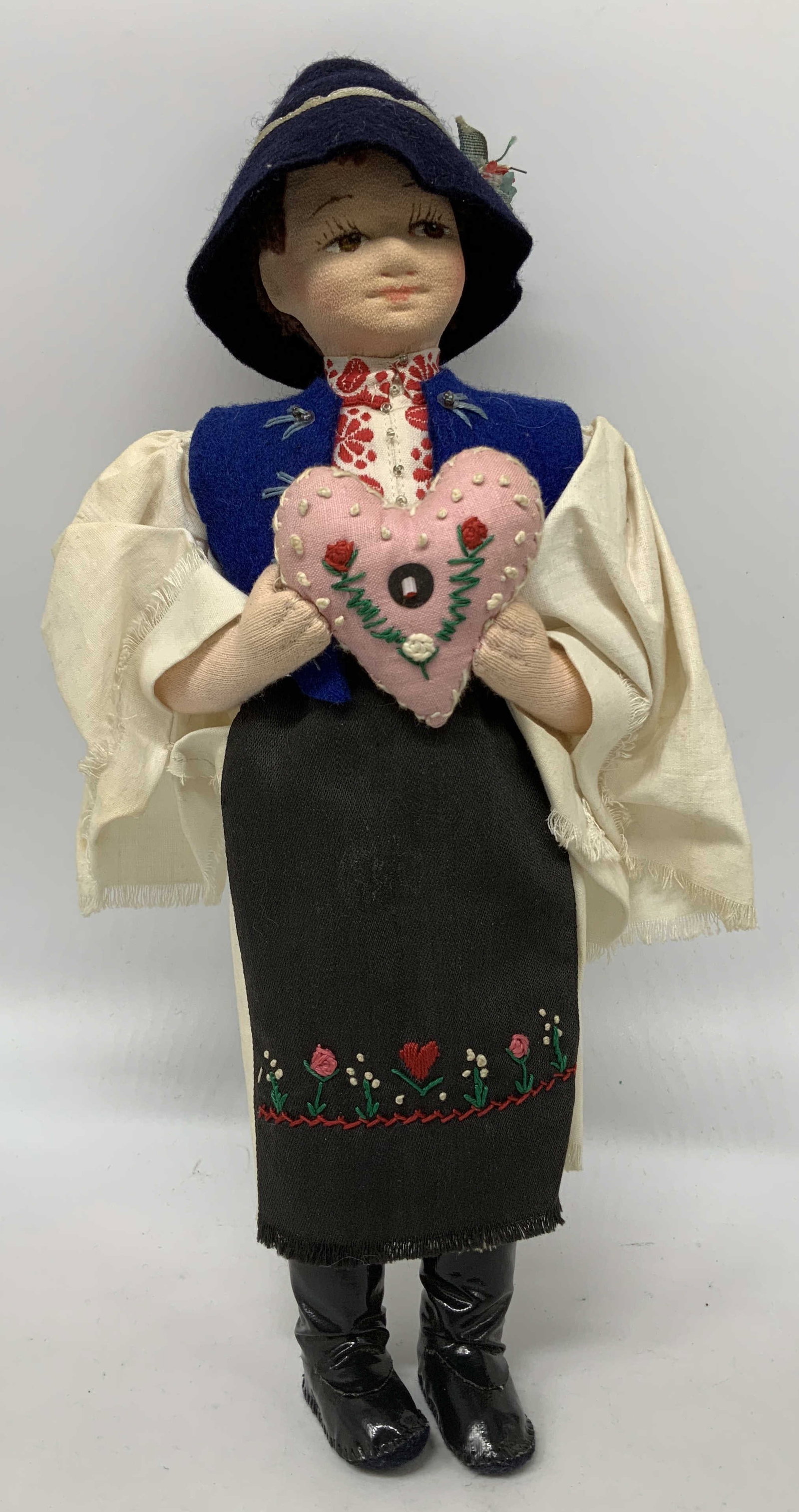 Hand Made Folk Art Doll W Embroidered Heart: Handcrafted folk art doll depicting a female figure wearing a blue vest, dark apron & white blouse. Hand embroidered details on clothing. She is holding a pink heart. Face is composite material. Surfa