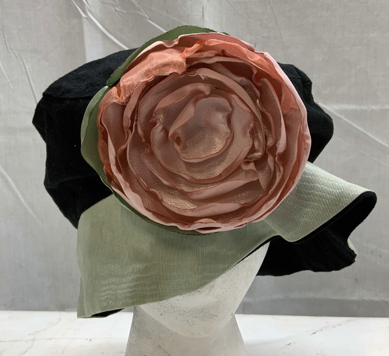 MESHUGENAH Black & Green Floral Hat: Vintage black & green hat w a large pink rose accent in center. Piece has a green brim. Surface wear consistent to age, use & exposure. Signed & marked. Reads, MESHUGENAH HAT COMPANY. Measurements: le