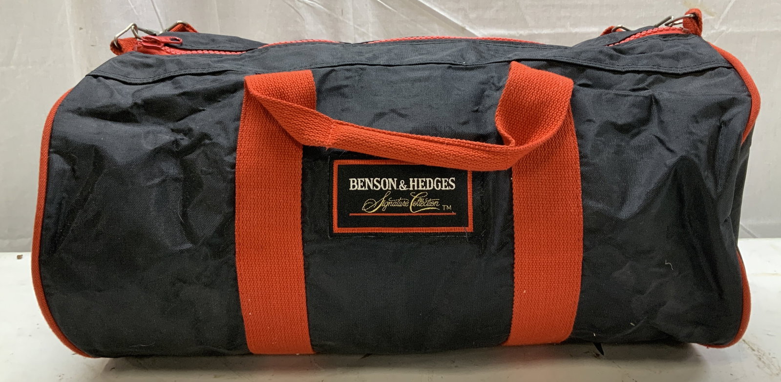 Vntg Benson & Hedges Cigarettes Promo Duffel Bag: Appears to be circa 1980s deadstock. Benson & Hedges promotional gym duffel bag. Black & red bag with detachable shoulder strap. Zipper closure. Signed & marked. Reads, BENSON & HEDGES SIGNATURE COLLE