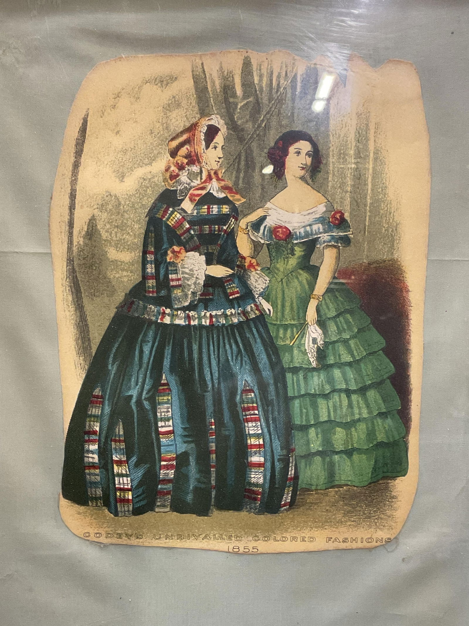 Framed 1855 Godeys Fashion Chromolithograph: Framed and mounted cut out Chromolithograph plate. Godeys Unrivaled Colored Fashions, 1855. Two women in period costume. Gilt wood frame. Not examined out of frame. Frame measures approx 12.75 x 9.75