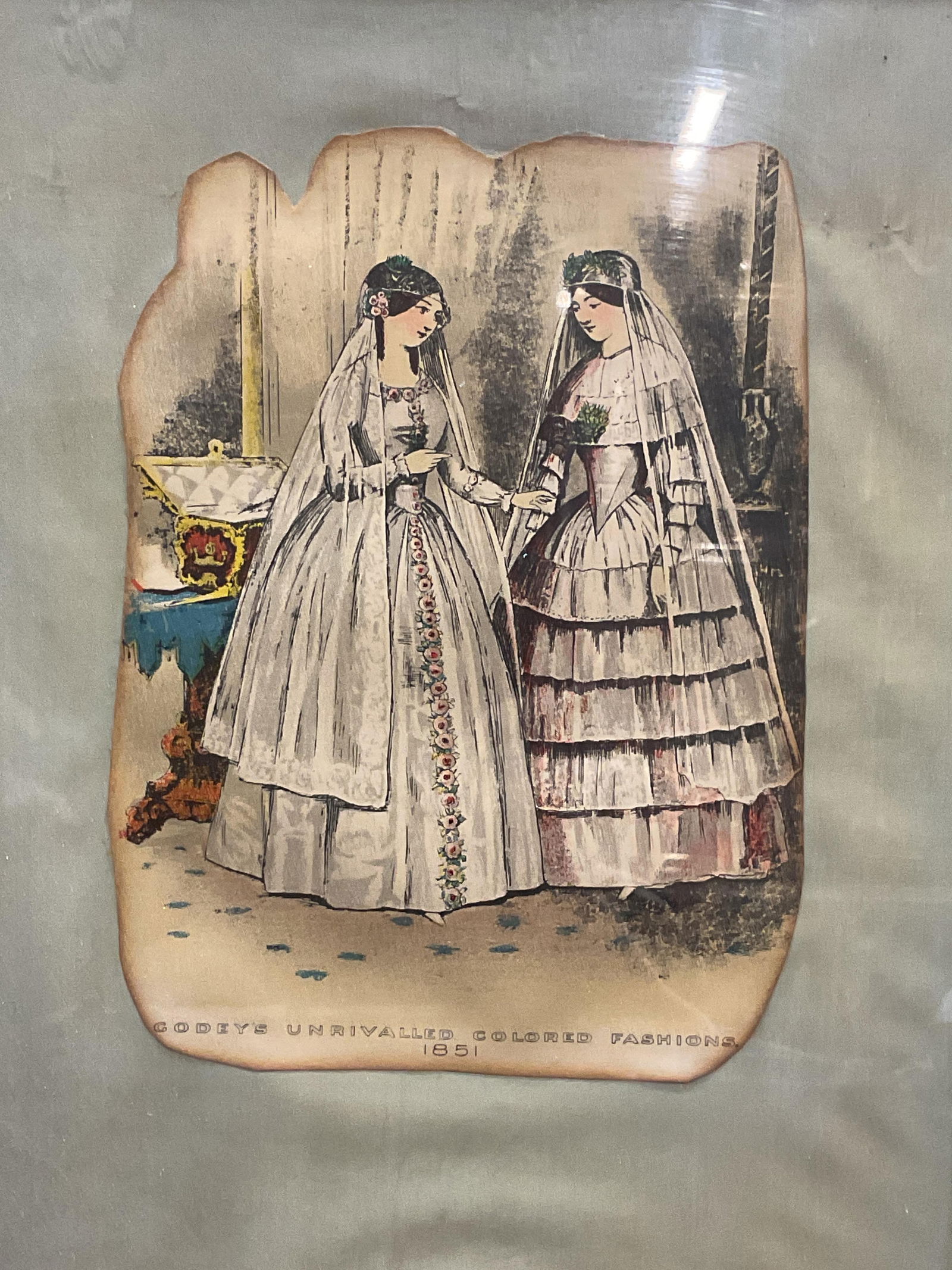 Framed 1851 Godeys Fashion Chromolithograph: Framed and mounted cut out Chromolithograph plate. Godeys Unrivaled Colored Fashions, 1851. Two women in period costume. Gilt wood frame. Not examined out of frame. Frame measures approx 12.75 x 9.75
