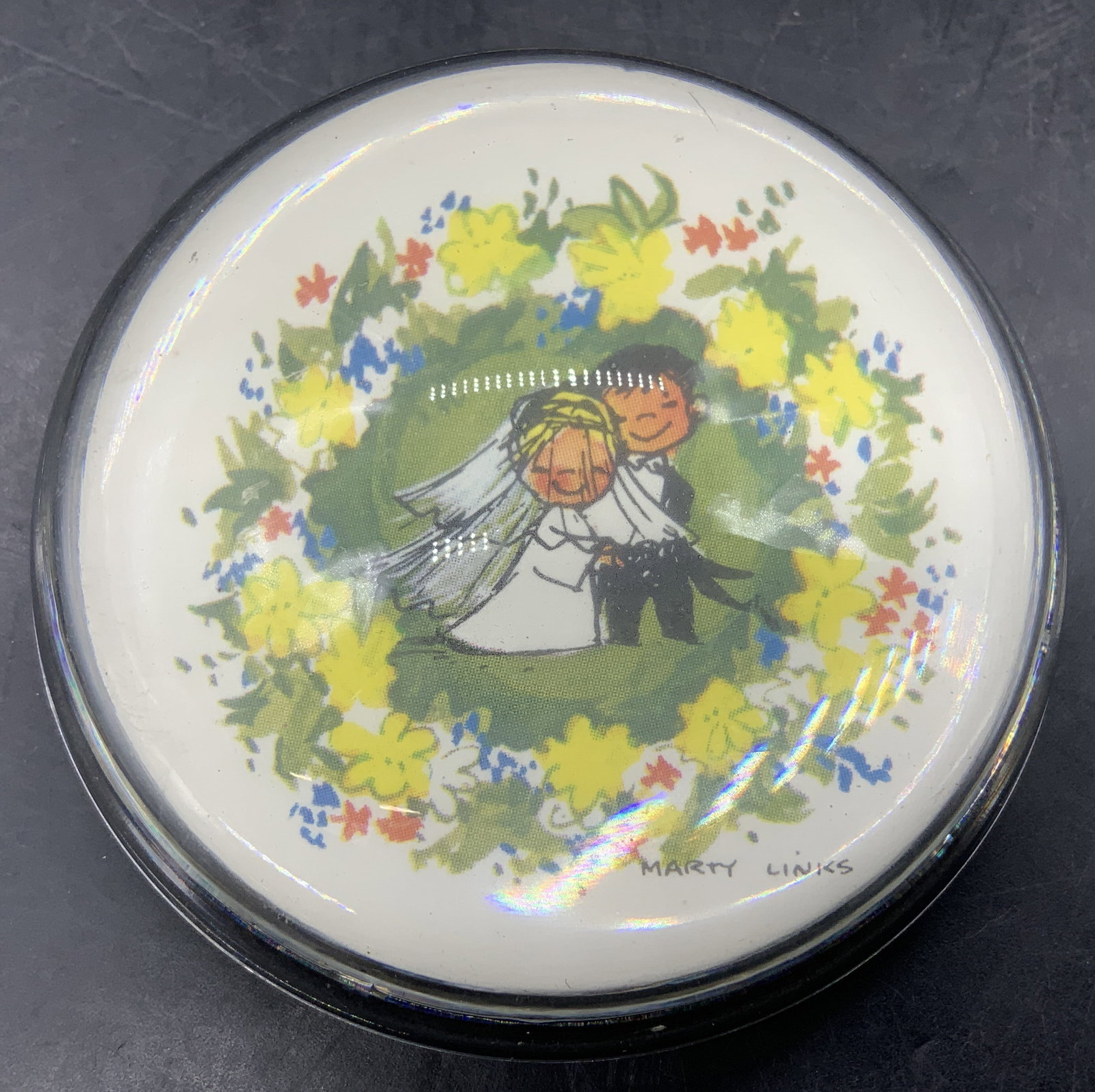 Marty Links Married Couple Paperweight: Paperweight featuring artwork by Marty Links, depicting a newly married couple surrounded by floral motifs. Appears to be acrylic. Surface wear consistent to age, use & exposure. Measurements: length
