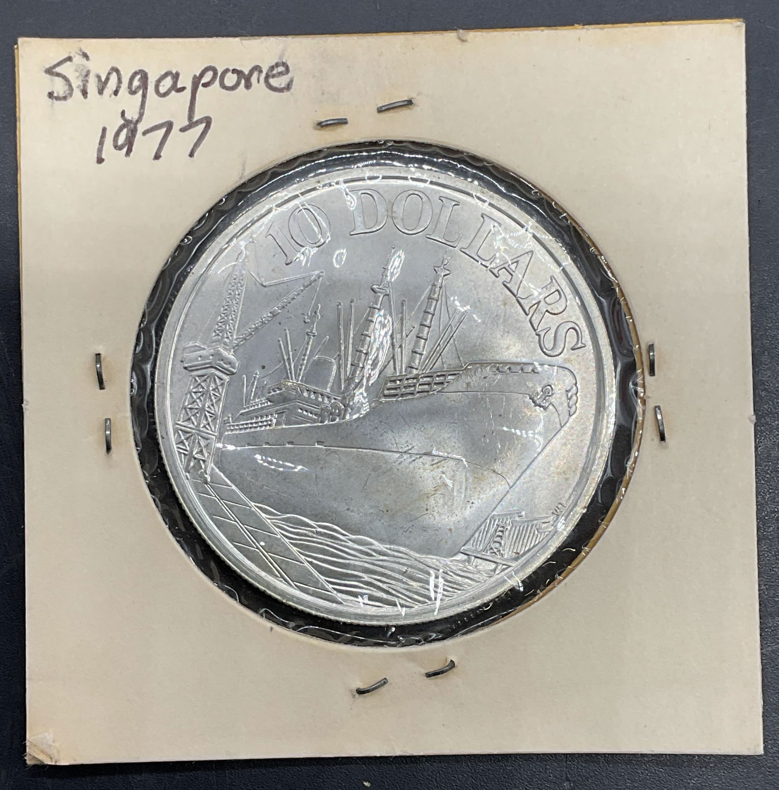 1977 Singapore 10 Dollar Coin, 500 Silver: 1977 Singapore 10 dollar coin. 500 silver. Reverse features flamingos container ship, obverse features coat of arms. Measures approx 1.5 inches wide. Held in protective cover, not examined out of cove