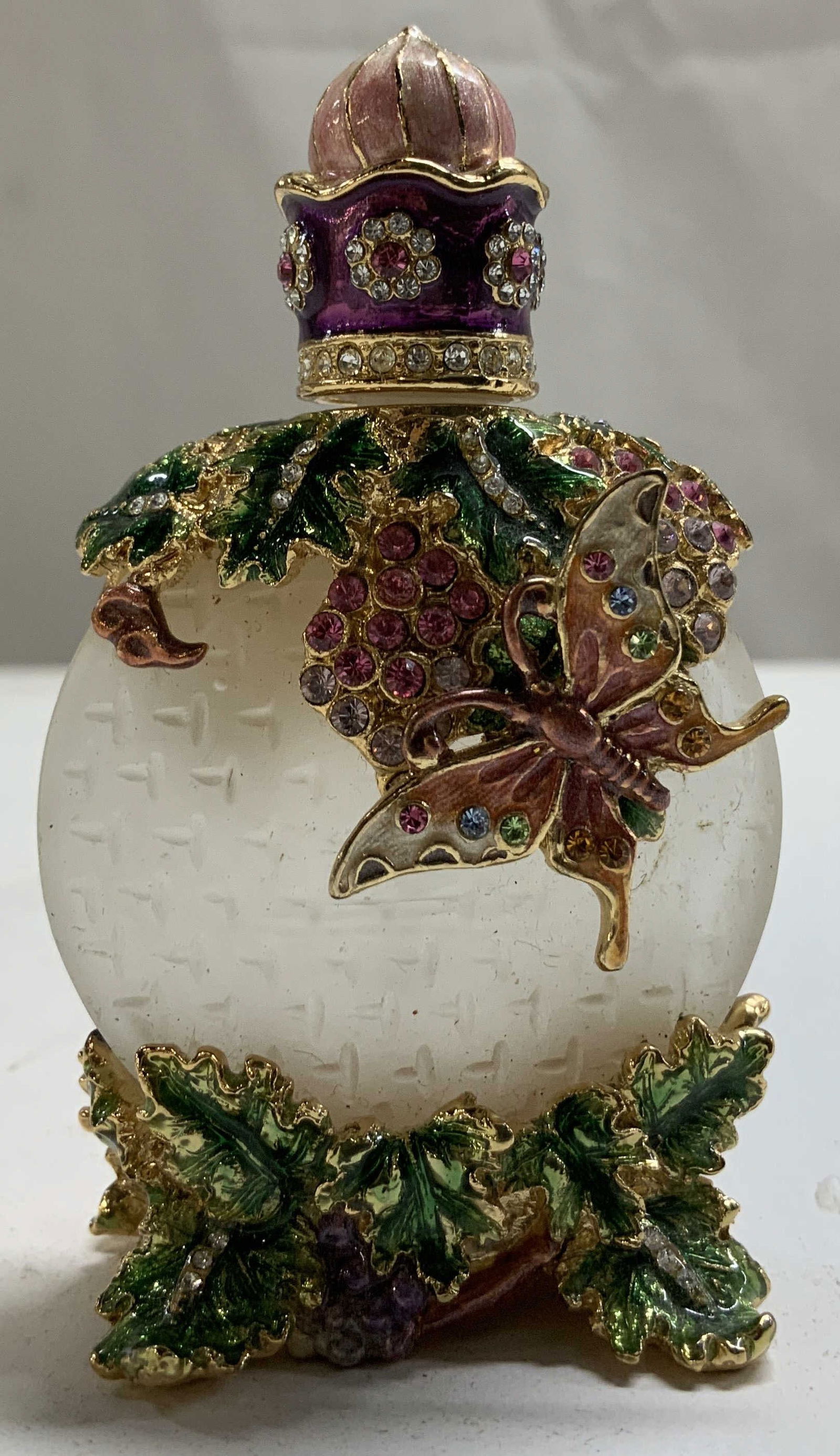 Bejeweled Enamel Butterfly & Leaf Perfume Bottle: Bejeweled enamel perfume bottle, featuring a butterfly & leaf design. Rhinestones accents throughout body. Shades of purple, pink, green, blue & yellow. Green leaves & grapes on base. Property of oper