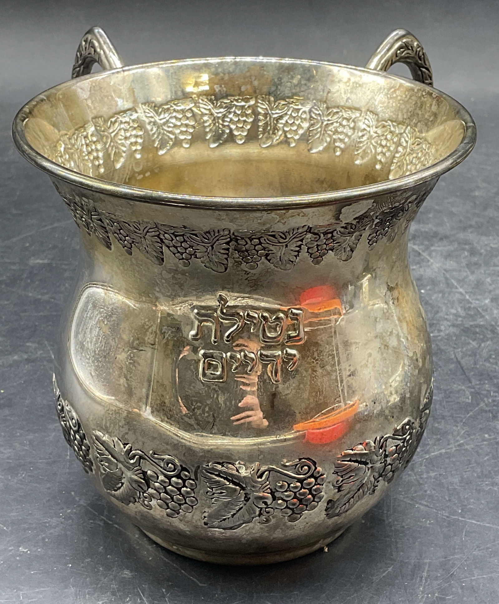 Netilat Yadayim Silver Plate Hebrew Washing Cup: Vintage silver plated Netilat Yadayim Hebrew washing cup. Judaica. Repousse grape vine pattern and Hebrew text. Double handled. Measures approx 4.5 x 6 x 4.75 inches. Wear including patina and scratch