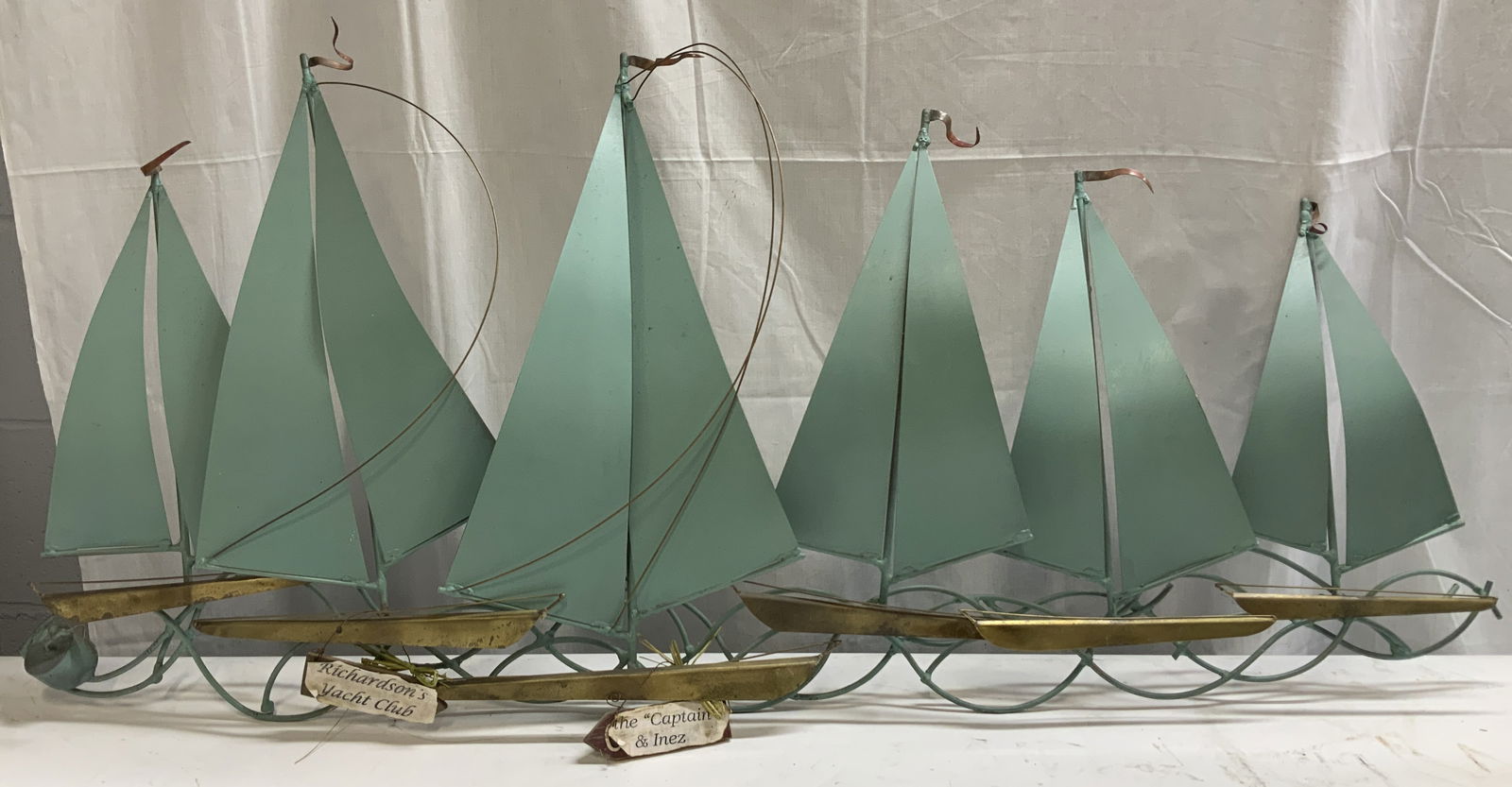 XL Stylized Sailboats Hanging Metal Wall Art 47in: Large decorative hanging metal wall art featuring a group of stylized sailboats with triangular green sails & brass toned hulls. Wire mast accents on sailboats. Base of hulls has name plates that read
