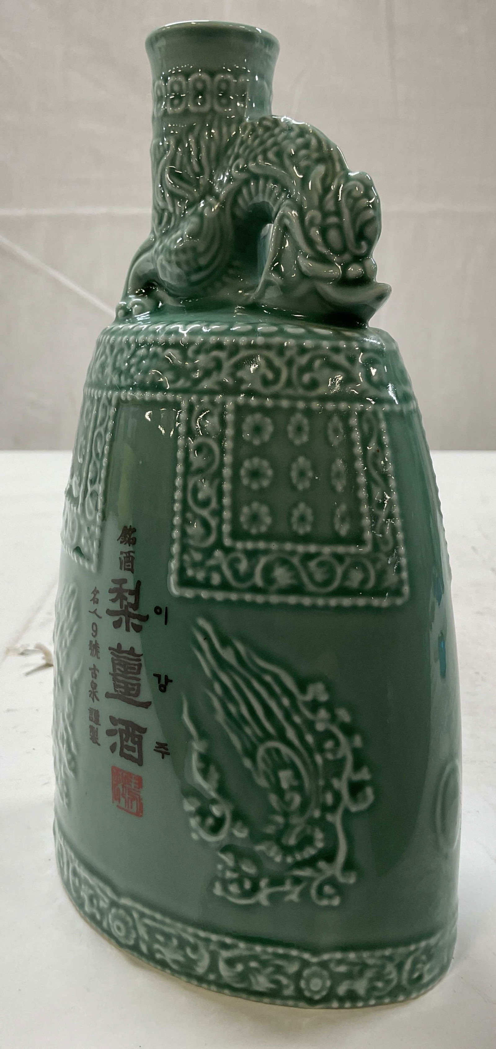 Asian Celadon Ceramic Jug With Dragon: Asian celadon ceramic jug with dragon. Raised floral motif and geometric patterns. Decorative dragon handle at the top. Signed on side and underside with Asian characters. Measures ht approx 8