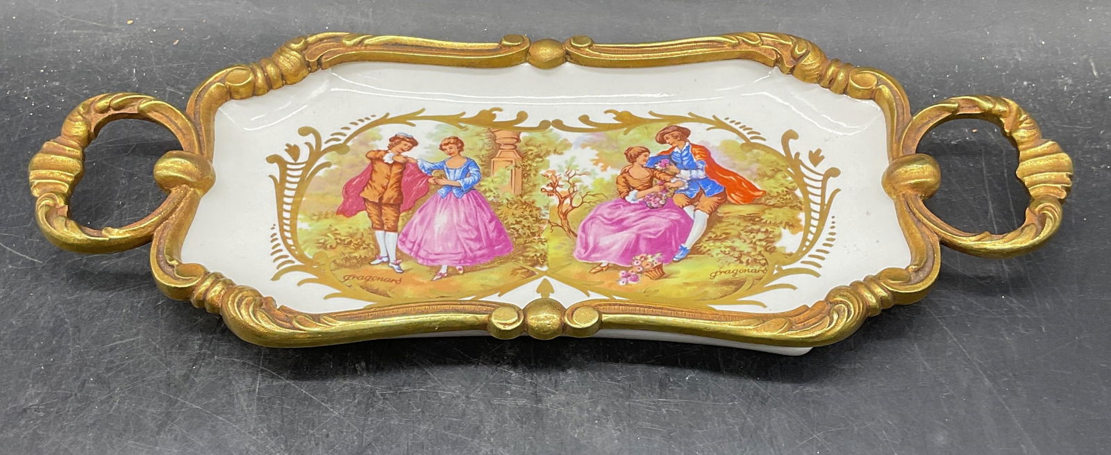 Signed Porcelain Fragonard Love Scene Tray, France: Signed porcelain tray with gilt brass handles and frame. Fragonard love scene motif. Signed with D inside of cartouche. Made in France. Measures approx 11.5 x 4.5 x 1 inches. Some wear including