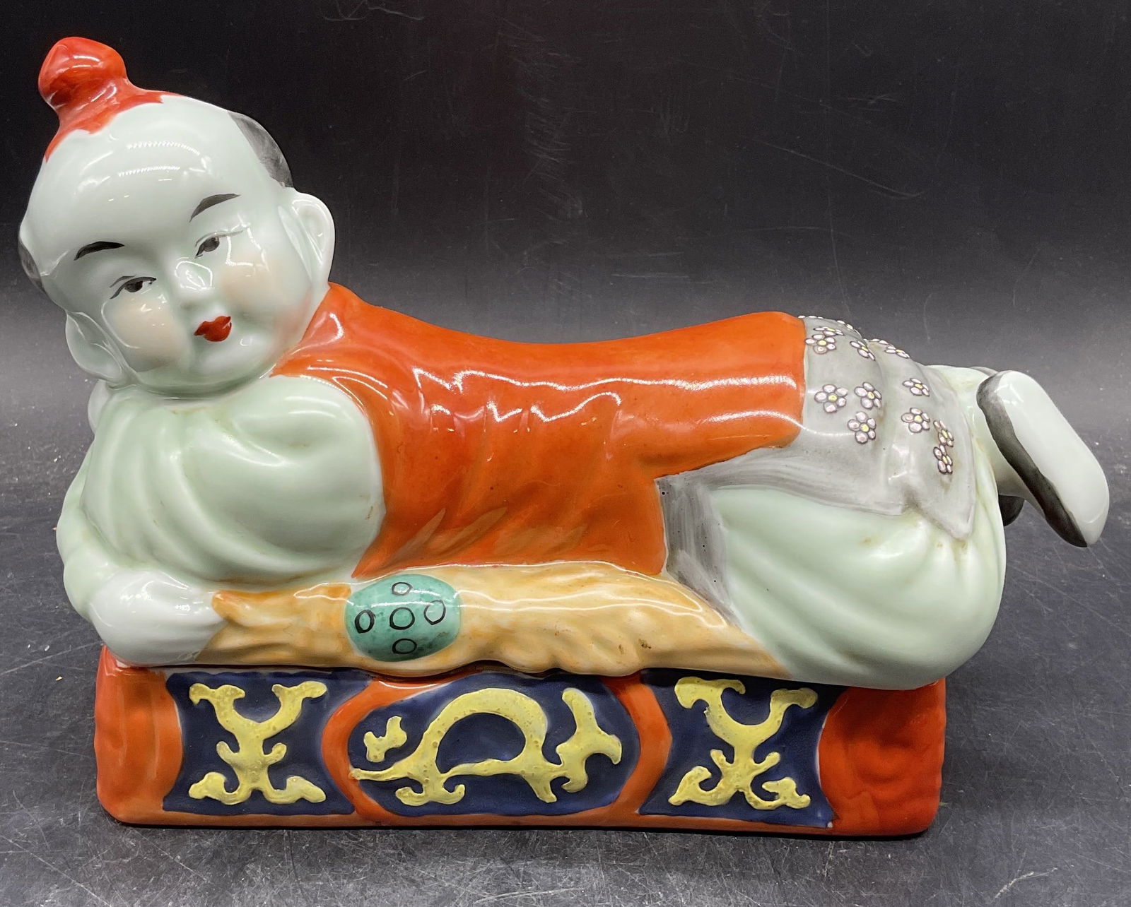 Chinoiserie Porcelain Pillow Figural Box: Chinoiserie porcelain pillow box. Figure of boy. Hand painted in orange and green. Measures approx 10.5 x 4.75 x 7.25 inches. Some wear including scratching. Chinoiserie box, porcelain box,