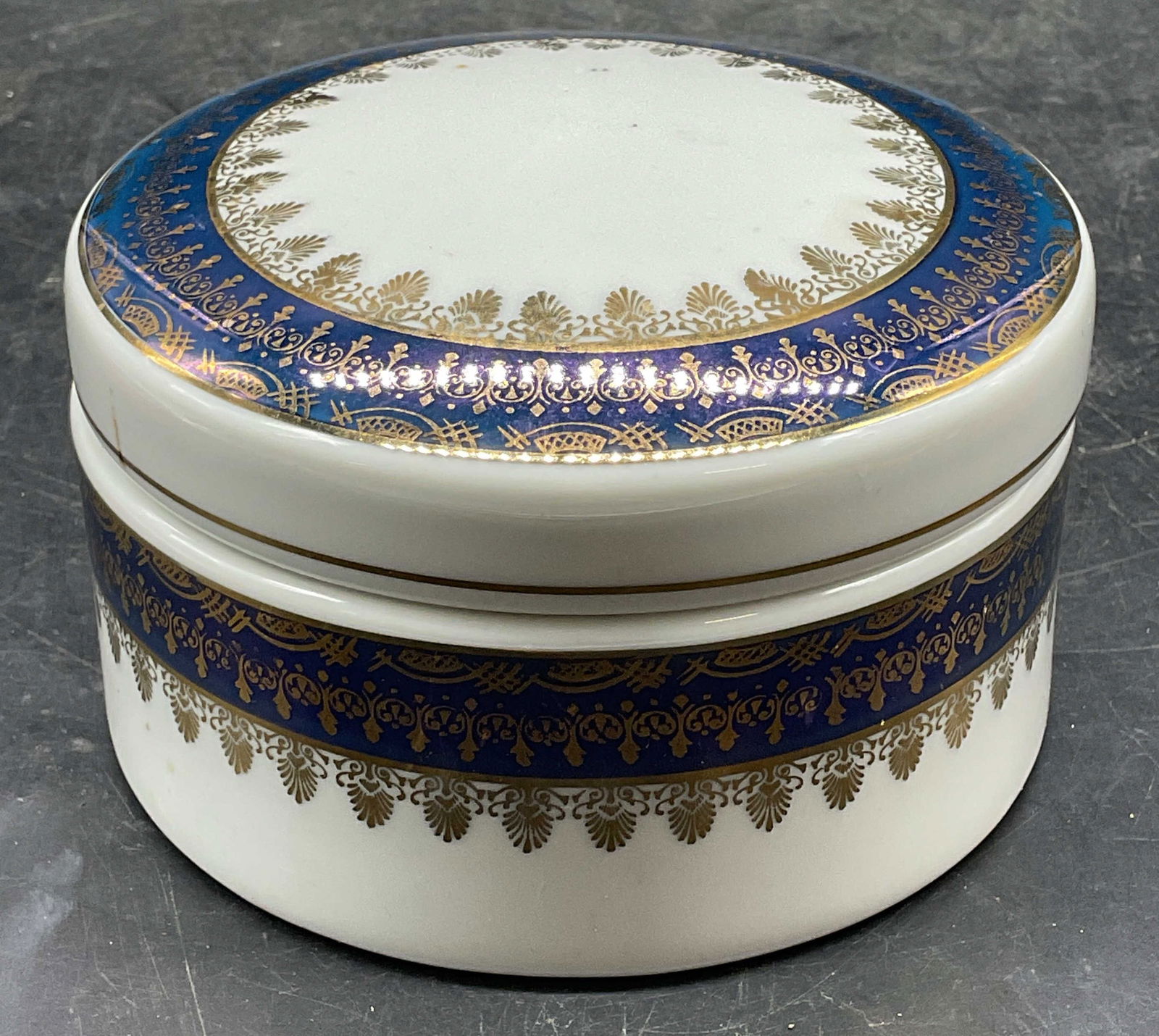 Pomerode Leart Round Gilt Porcelain Box, Brazil: Pomerode Leart round porcelain lidded box. Cobalt blue and gilt border on white. Ornate scroll design. Measures approx 5 x 2.75 inches. Signed. Made in Brazil. Some scratching and marks. Pomerode