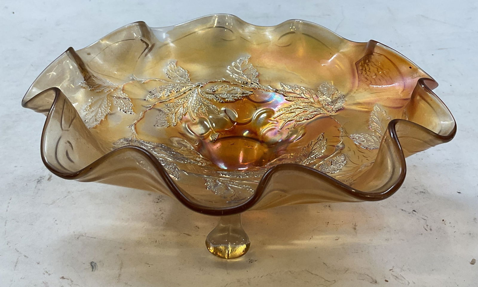 Vintage Scalloped Iridescent Glass Footed Bowl: Vintage scalloped iridescent carnival glass footed bowl. Scalloped and ruffled edge. Iridescent orange finish. Embossed leaf and cherry pattern. Three splayed feet on underside. Measures ht approx 3 i