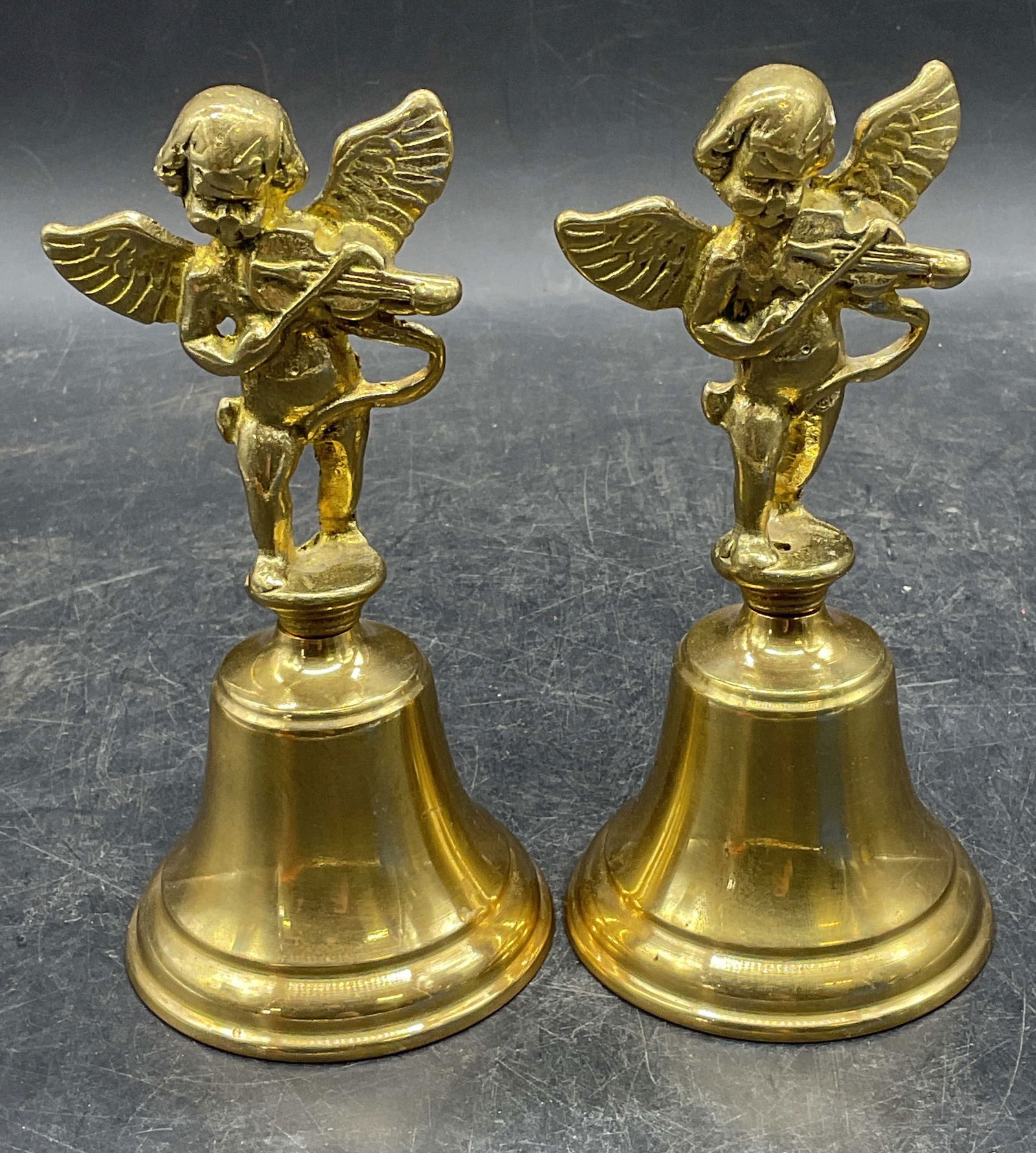 Pair Brass Cherub Musician Hand Bells: Pair of brass hand bells with cherub playing violin motif handles. Measure approx 3 x 6 inches. Some scratching. Brass bell, hand bells, cherub bell, home decor, home accessories, table decor, table a