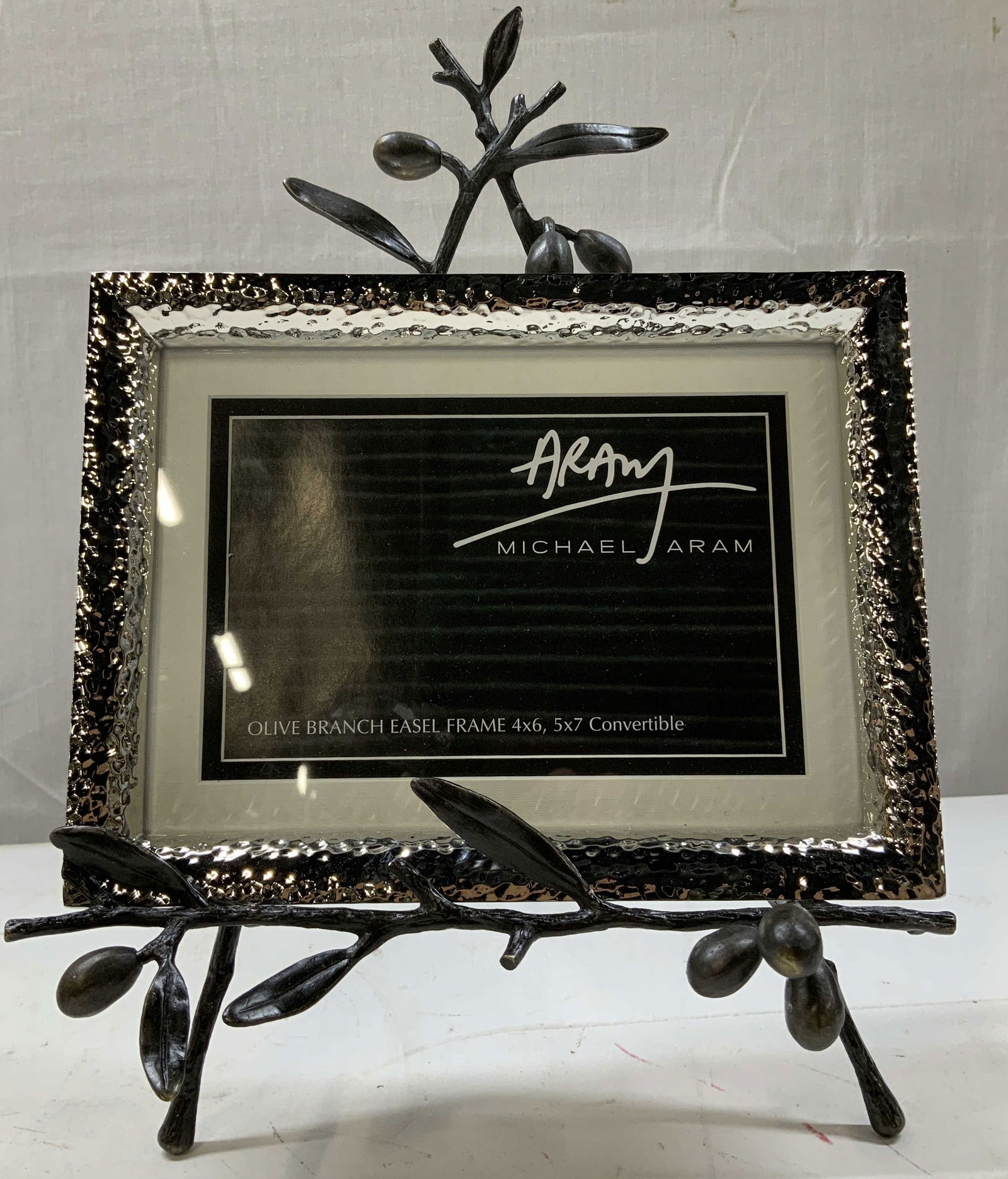 Michael Aram Olive Branch Easel Convertible Frame: Easel features a detailed design of intertwining olive branches & leaves. Piece is made of cast metal. Frame has a nickel plated finish. Signed & marked. Reads, Michael Aram. Measurements: length 7.8i