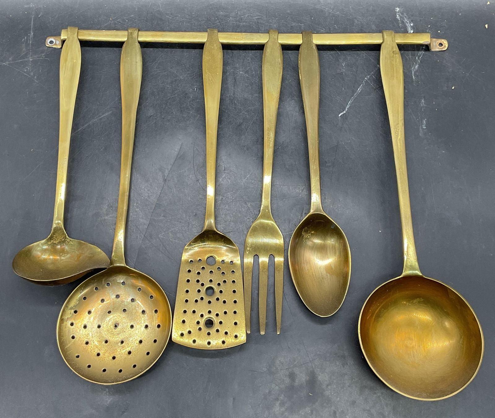 Set 7 Vintage Brass Kitchen Utensils & Rack: Set of 7 vintage brass kitchen utensils with hanging rack. Includes fork, spoon, slotted spatula, skimmer, and 2 ladles. Largest piece approx 13.75 inches long. Rack approx 16.5 inches wide. Wear incl