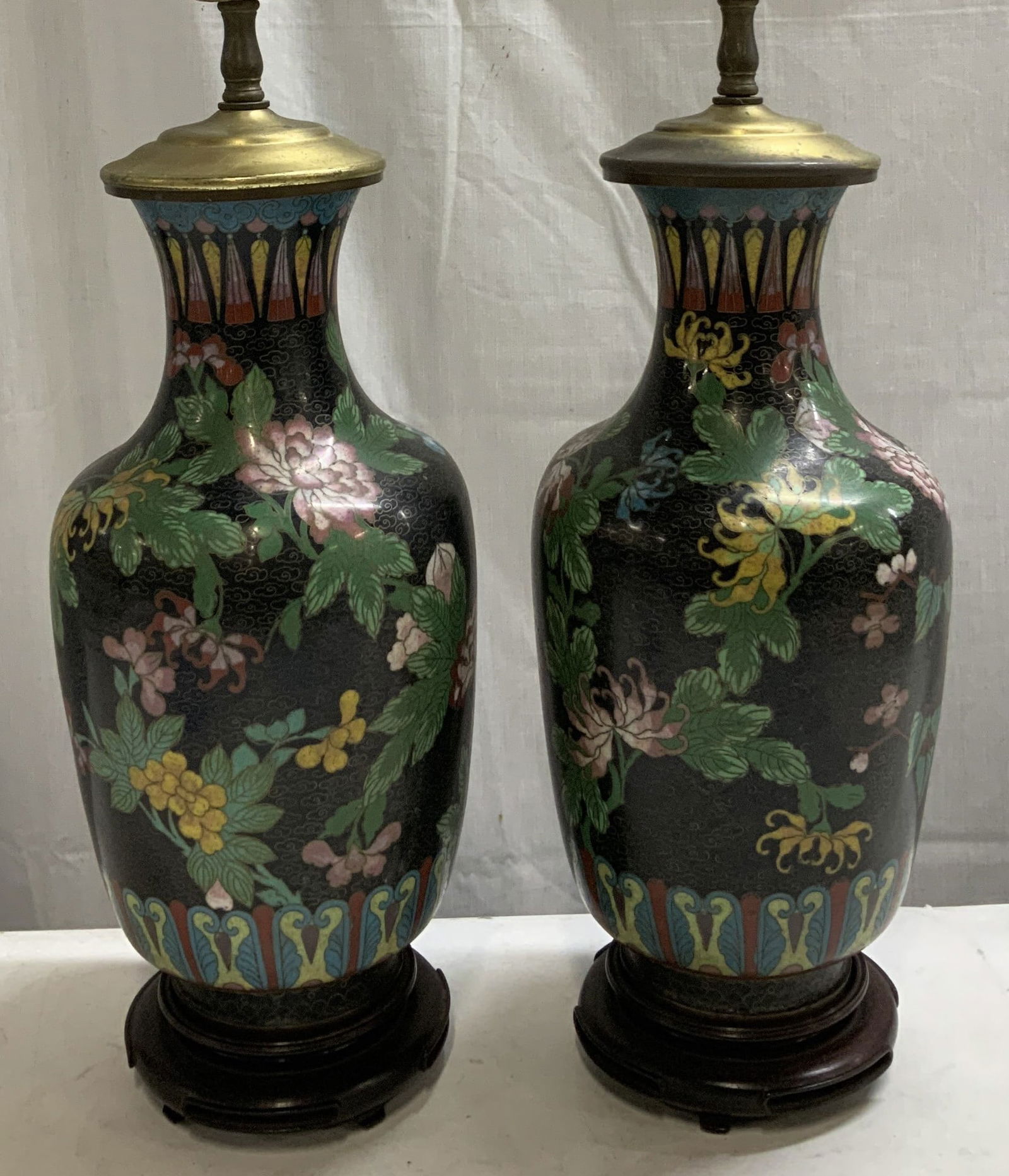 Pair Vtg Cloisonne Enamel Floral Table Lamps: Lot includes, a pair of vintage cloisonne enamel table lamps. Lamps feature a black background with floral detailing. Shades of green, yellow, red & pink. Sits on a round wooden base. Table lamps have