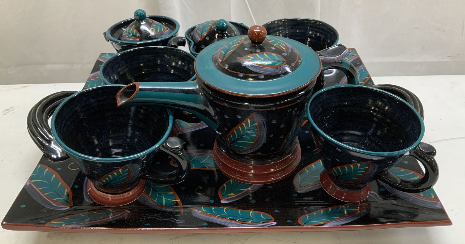 Set 8 Sgnd Page Thorbeck Studio Pottery Teapot Set: Lot includes, 1 lidded sugar bowl, 1 creamer, 4 teacups, 1 teapot & serving platter to hold teapot set. Pieces feature hand painted leaf accents. Shades of blue, red, & purple. Black toned.