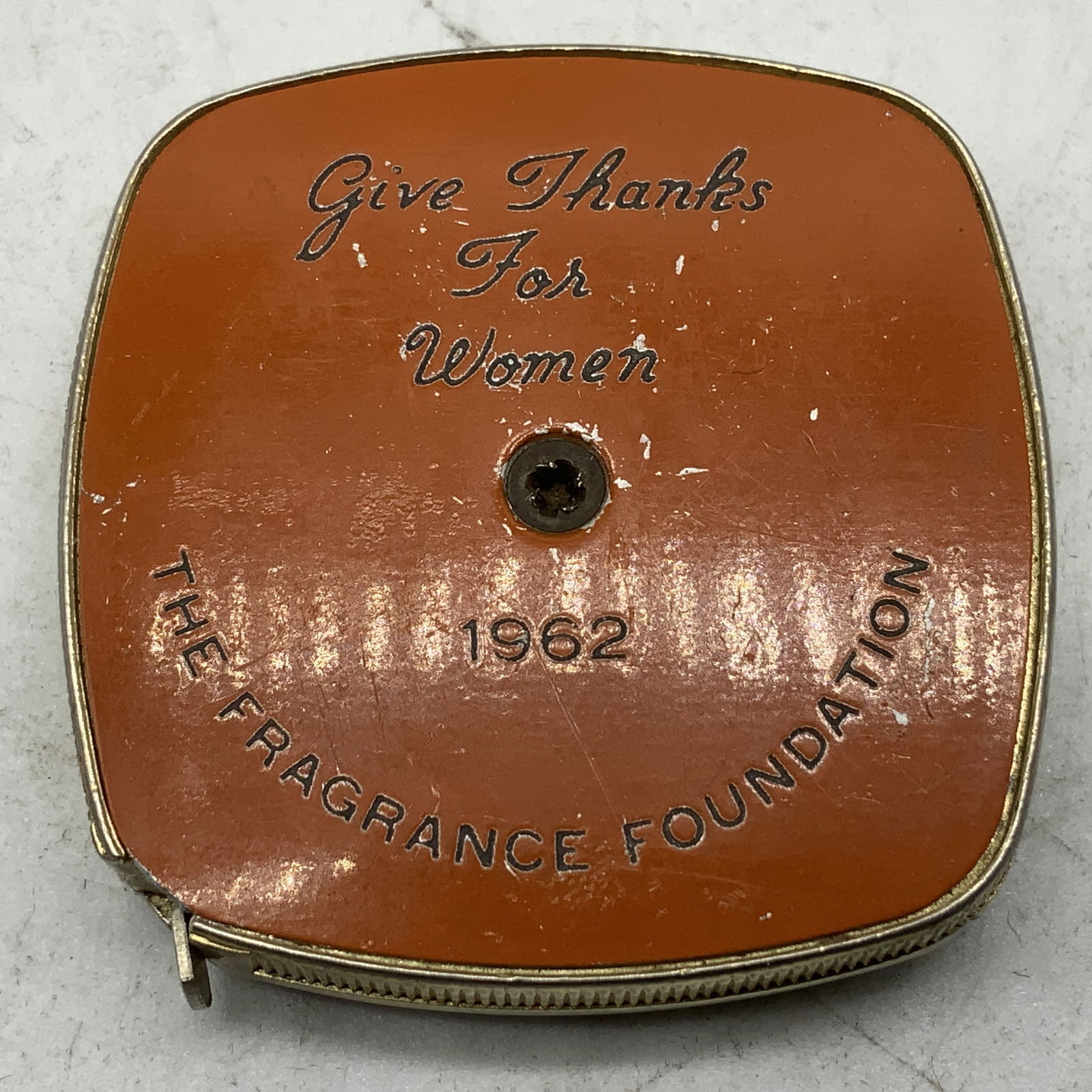 1962 The Fragrance Foundation Tape Measure USA: Retractable tape commemorating Give Thanks For Woman from the Fragrance Foundation in 1962. Tape measure features an orange toned front & silver toned back. Surface wear consistent to age, use &