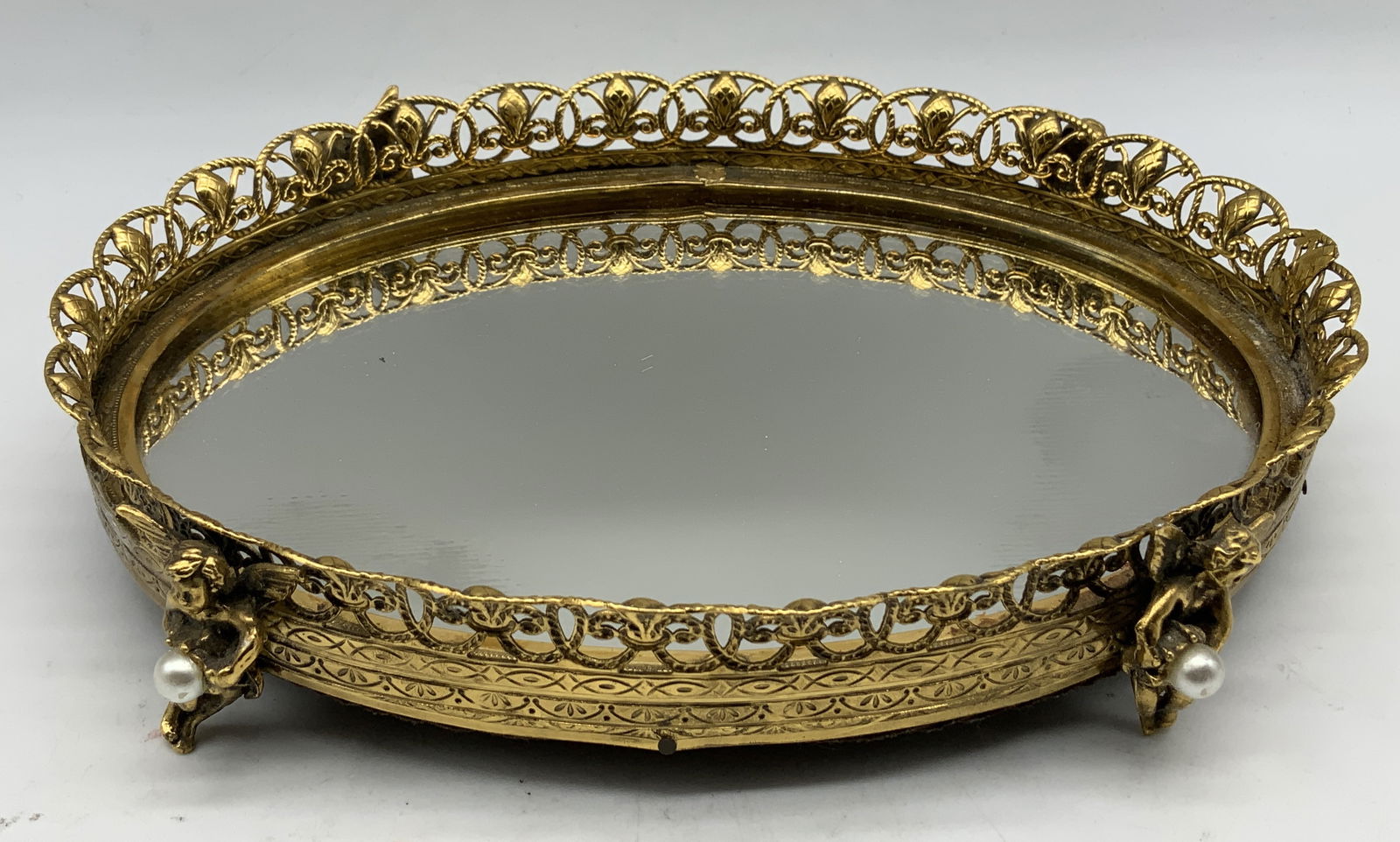 Vtg Stylebuilt Brass Cherub Mirror Dresser Tray: Vintage oval dresser tray features a brass ornate frame & raised on 4 legs. Legs are shaped as cherub figures holding faux pearls. Mirrored glass base. Surface wear consistent to age, use & exposure.