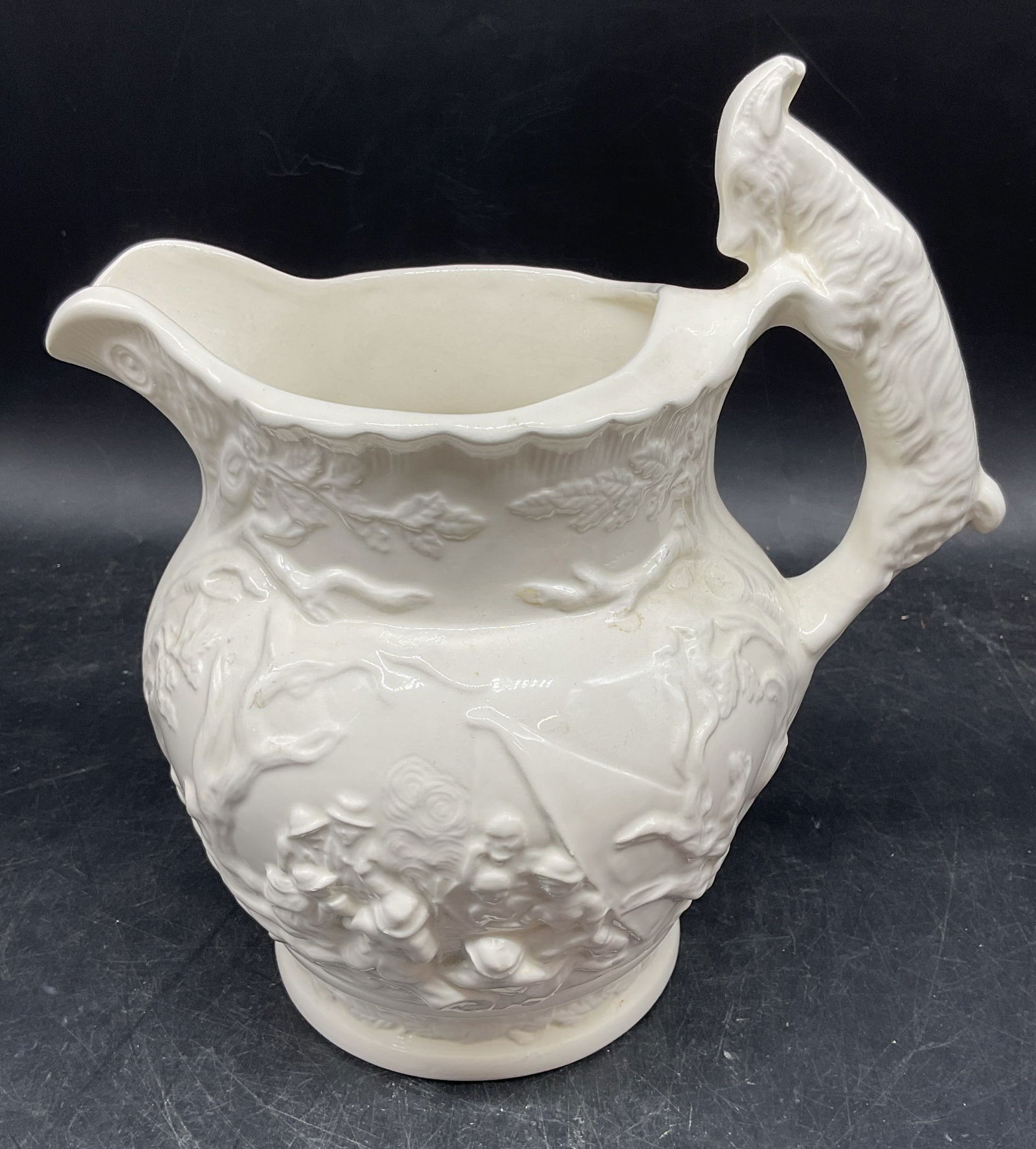 Signed White Ceramic Hunt Scene Pitcher: Signed white ceramic relief pitcher with hunting figural scene. Ram motif handle. Measures approx. 8 x 6 x 8.5 inches. Signed with makers mark. Wear including scratching. White pitcher, ceramic pitche