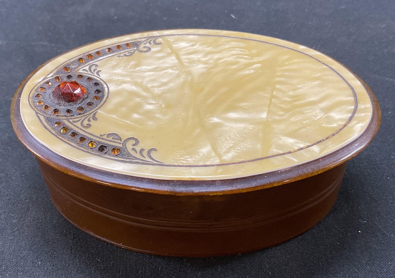 Vintage Art Deco Celluloid Lidded Powder Box: Vintage Art Deco celluloid lidded powder box. Amber colored oval shaped celluloid base. Cream colored mother of pearl style inlaid lid. Amber colored rhinestones. Ornate scroll motifs. Art Deco style.