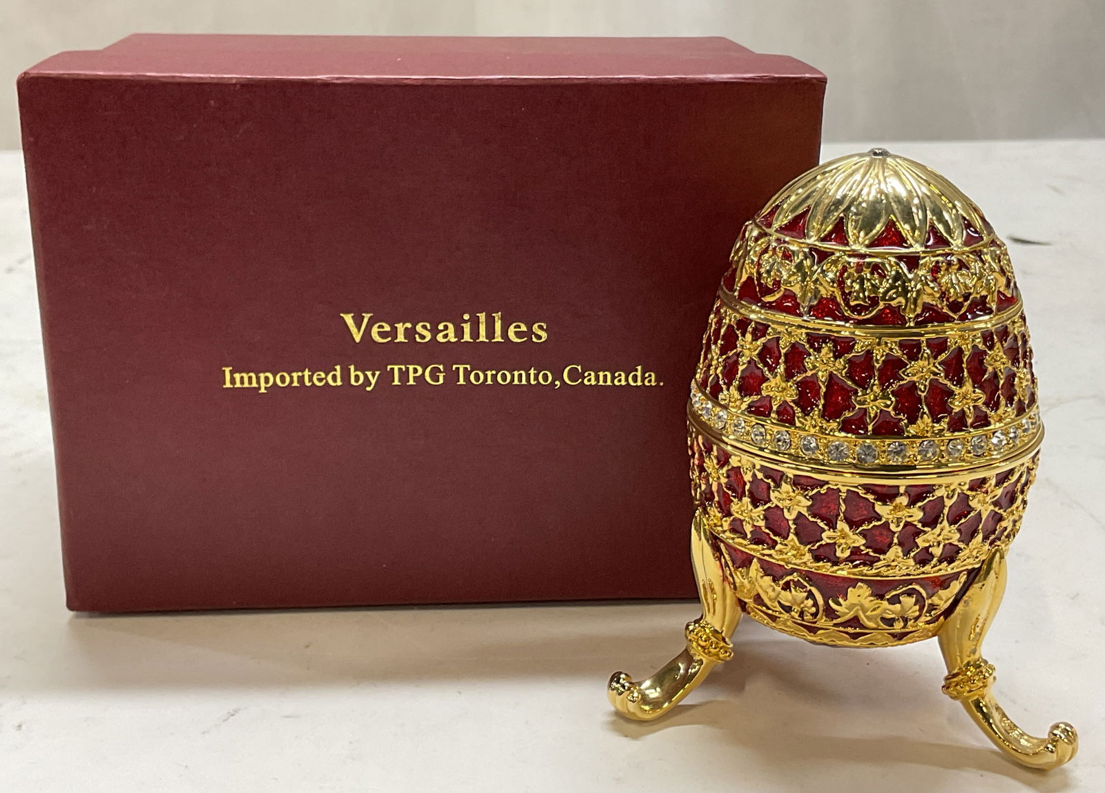 Versailles Egg Shaped Footed Trinket Box W Box: Versailles ruby red egg shaped hinged footed trinket box. Comes in original box. Egg shaped box, Faberge egg style. Sticker adhered to underside reads Versailles. Ruby red enameled background with gil