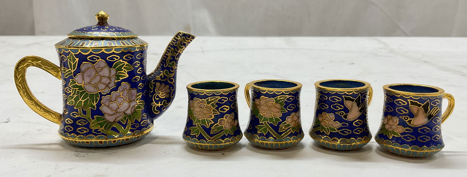 Set 6 Chinese Cloisonne Miniature Tea Set: 6 piece Chinese cloisonne miniature tea set. Includes tea pot with lid and 4 teacups. Enameled brass. Gilt decorations. Enameled floral and bird motifs. Colors consist of blue, green, and pink. Larges